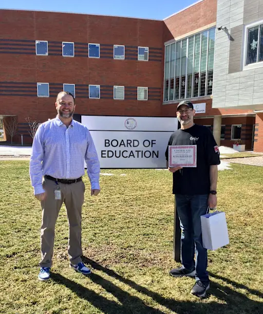 Two men stand outside a building with a "BOARD OF EDUCATION" sign, one holding a certificate.