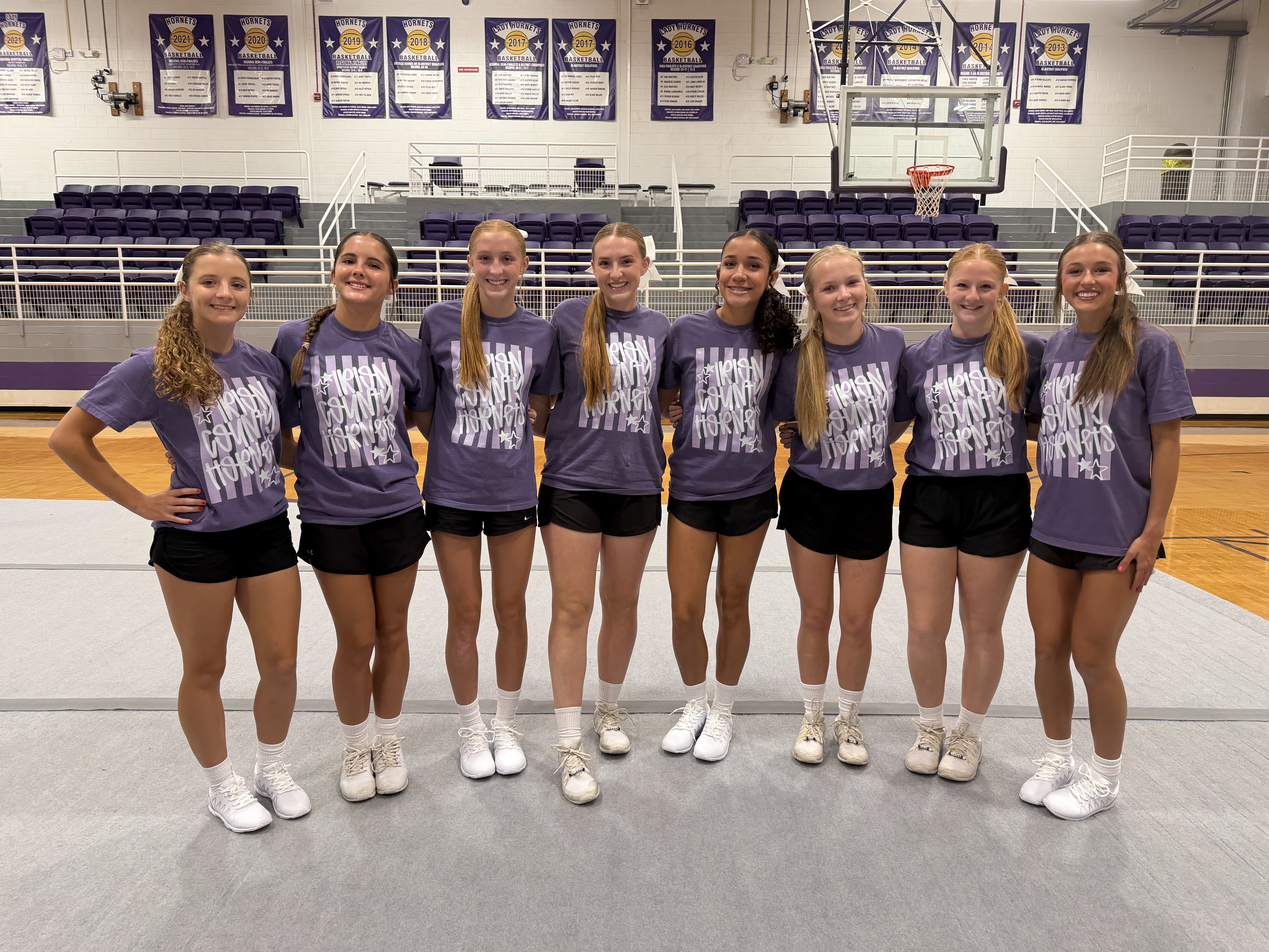 A group of eight young women in matching purple t-shirts and black shorts stand together in a gymnasium.