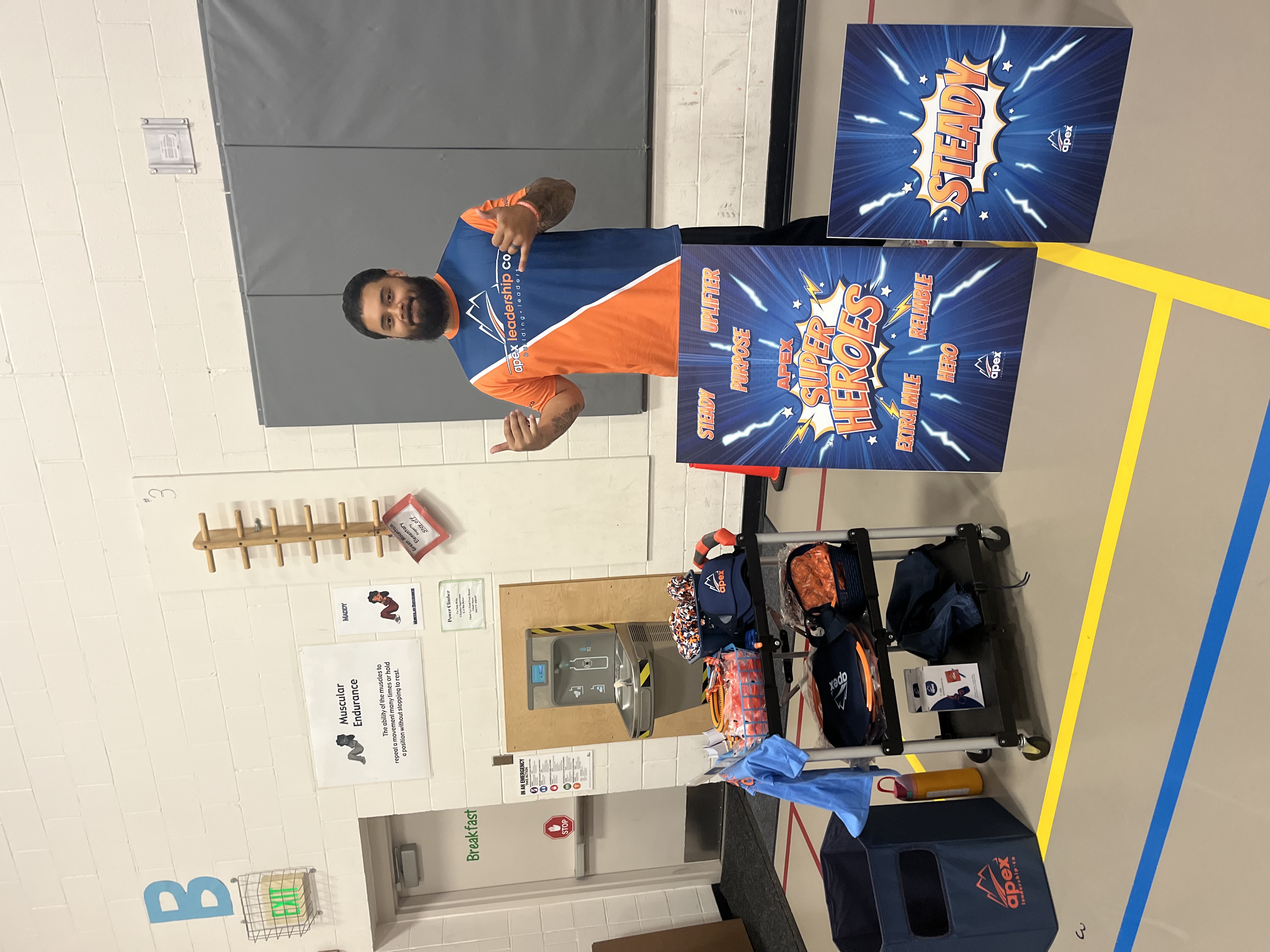 A smiling man stands in a gymnasium next to promotional signs for Apex Leadership Co. and a cart of gear.