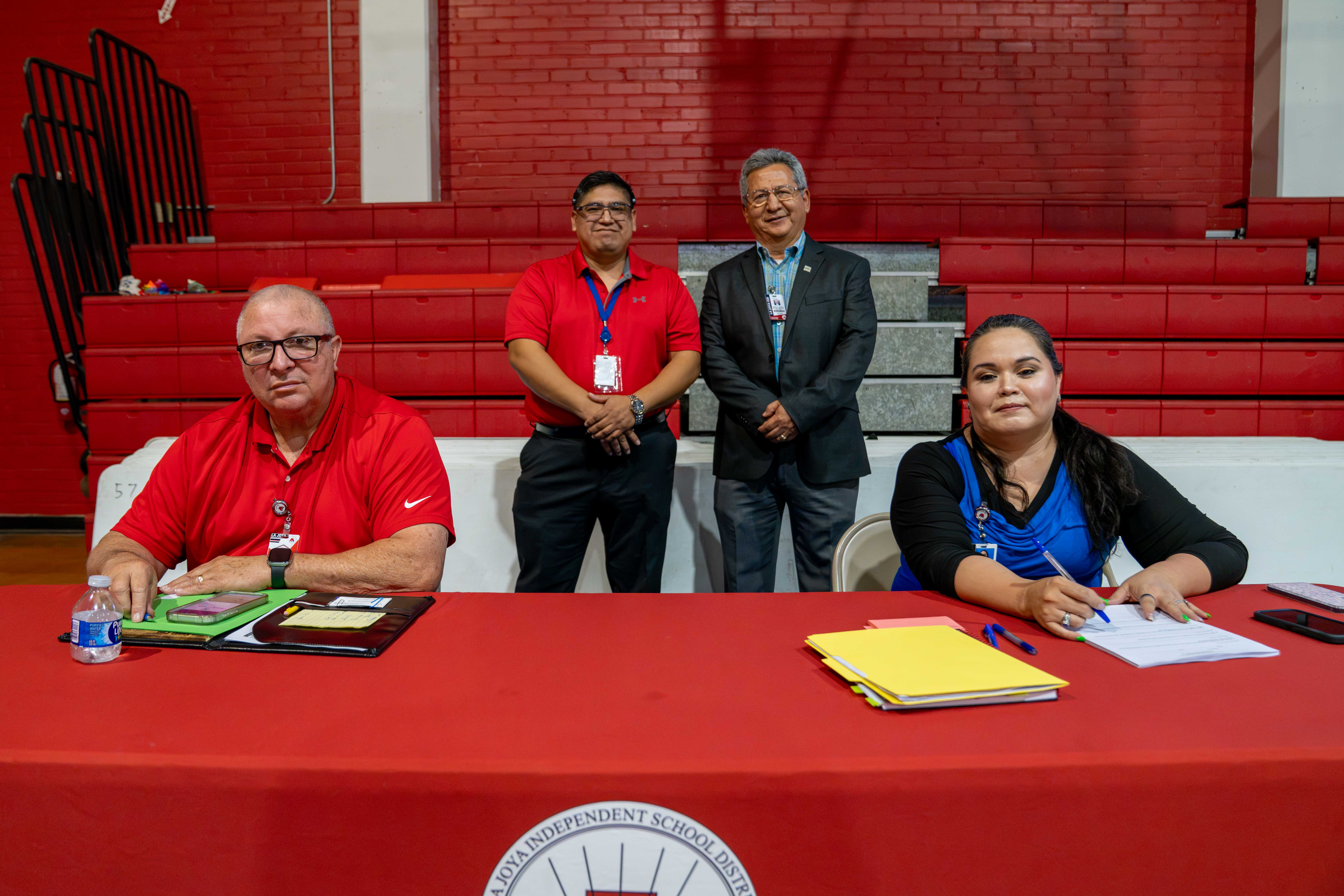 Four people pose behind a red table inside a gymnasium with red bleachers.