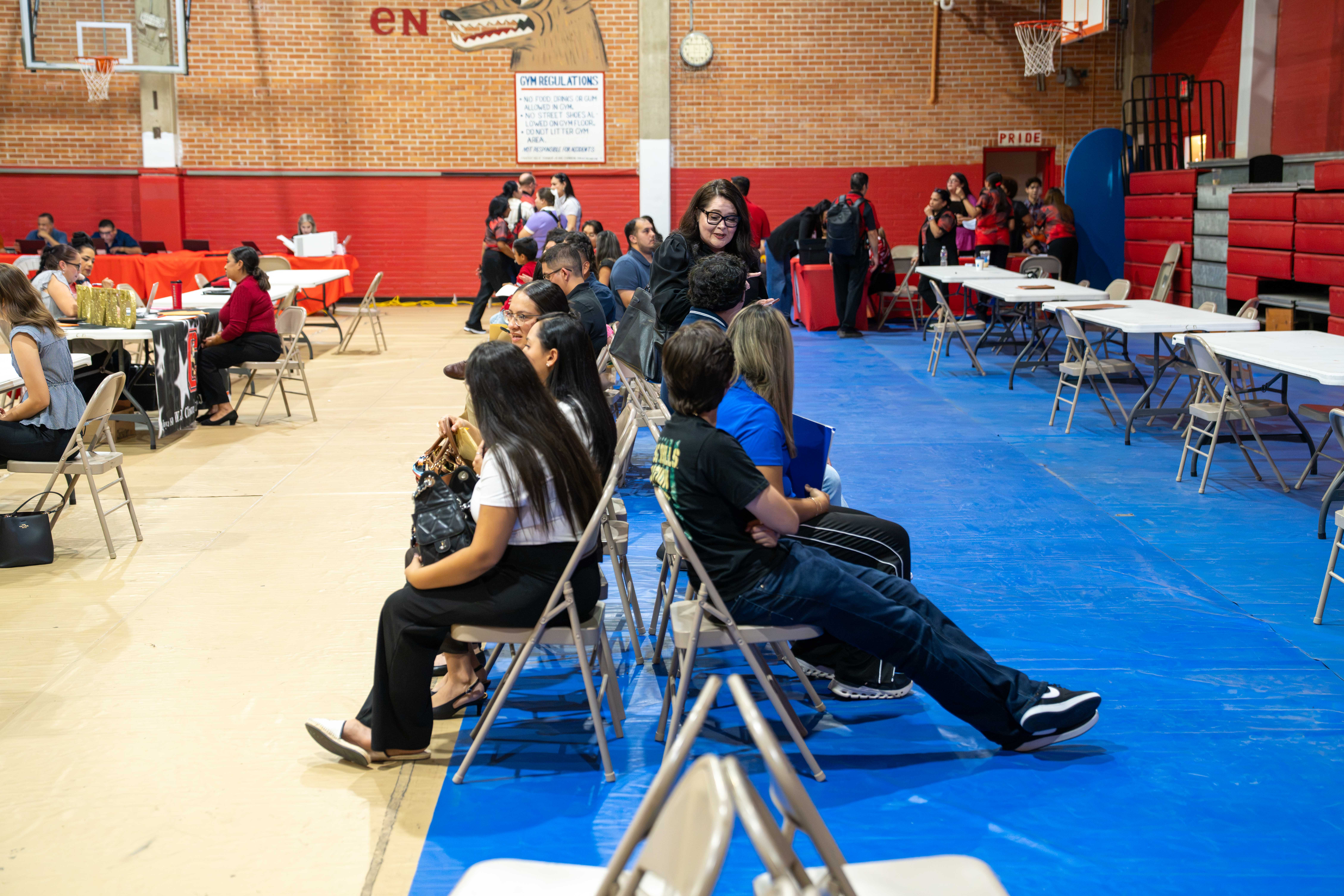 People sit in rows of folding chairs inside a gymnasium, attending a community or school event.