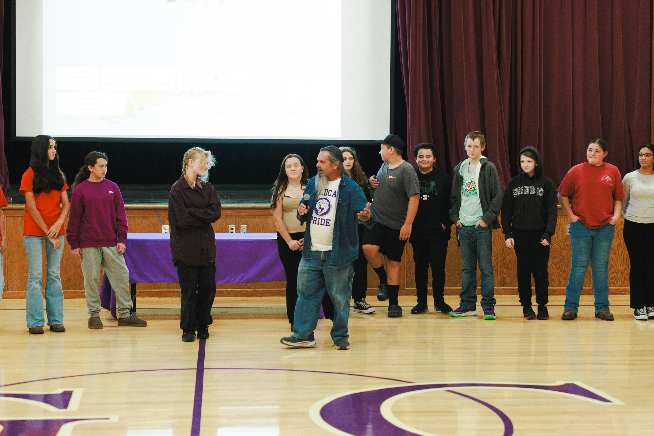 A speaker holds a microphone while addressing a group of young people standing on a school gymnasium stage.