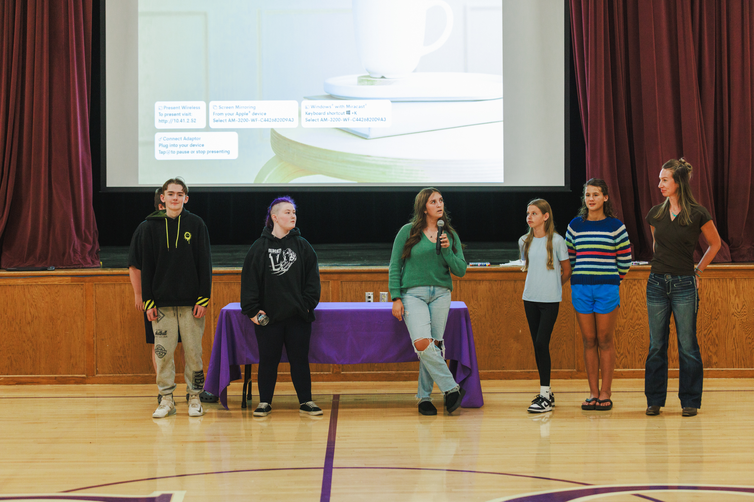 A group of students and a teacher stand on a gymnasium stage during a presentation.