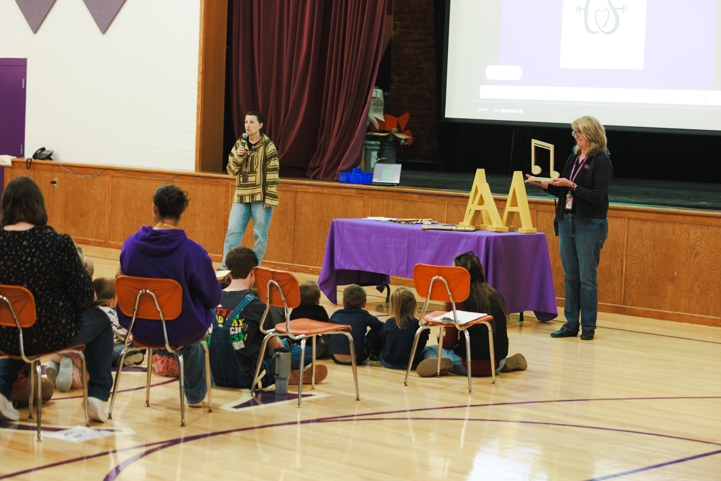 A speaker holds a microphone on a stage while another person displays a musical note cutout, presenting to an audience seated in a gymnasium.
