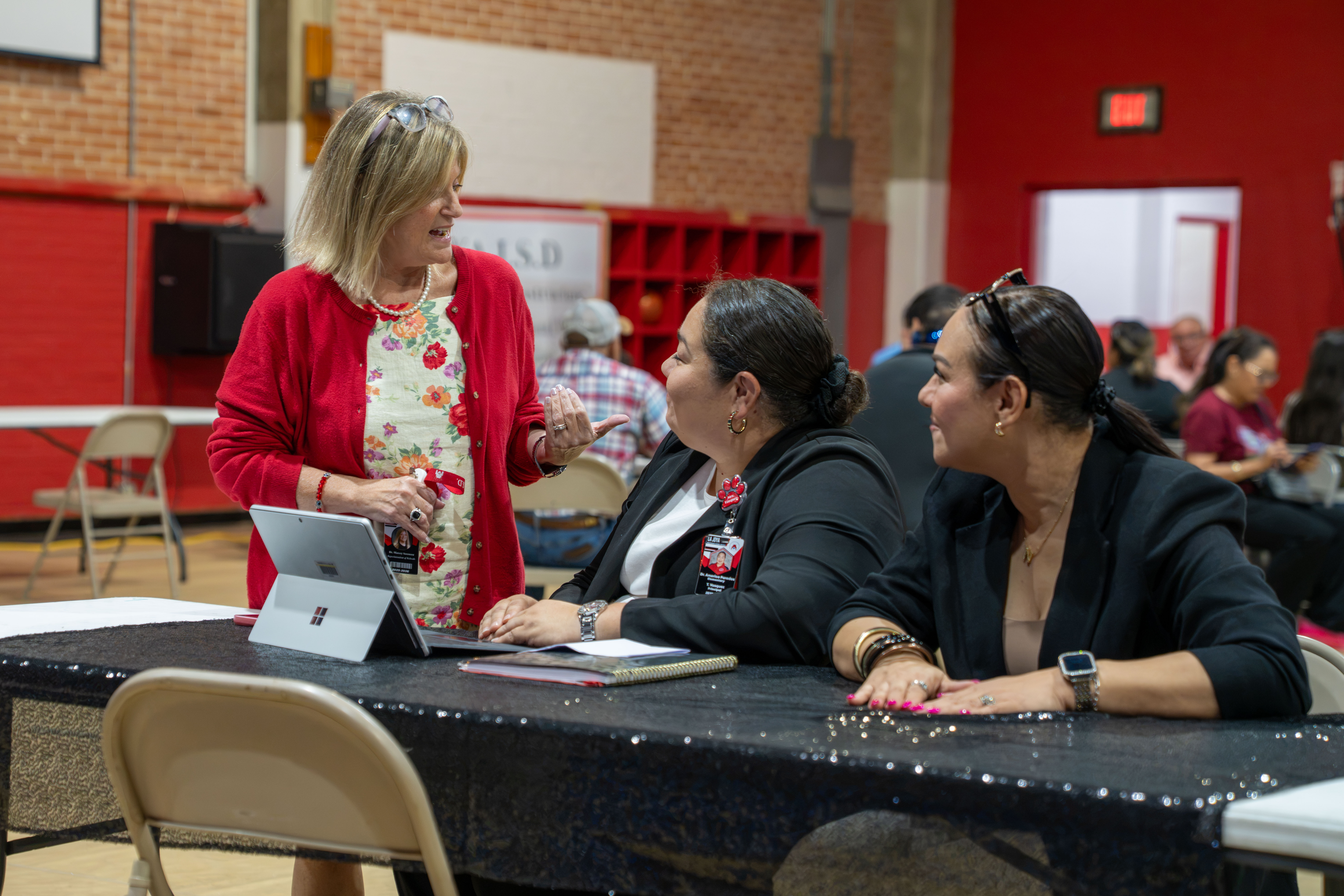 Three women engage in a discussion at a table, with a laptop and notebooks present.