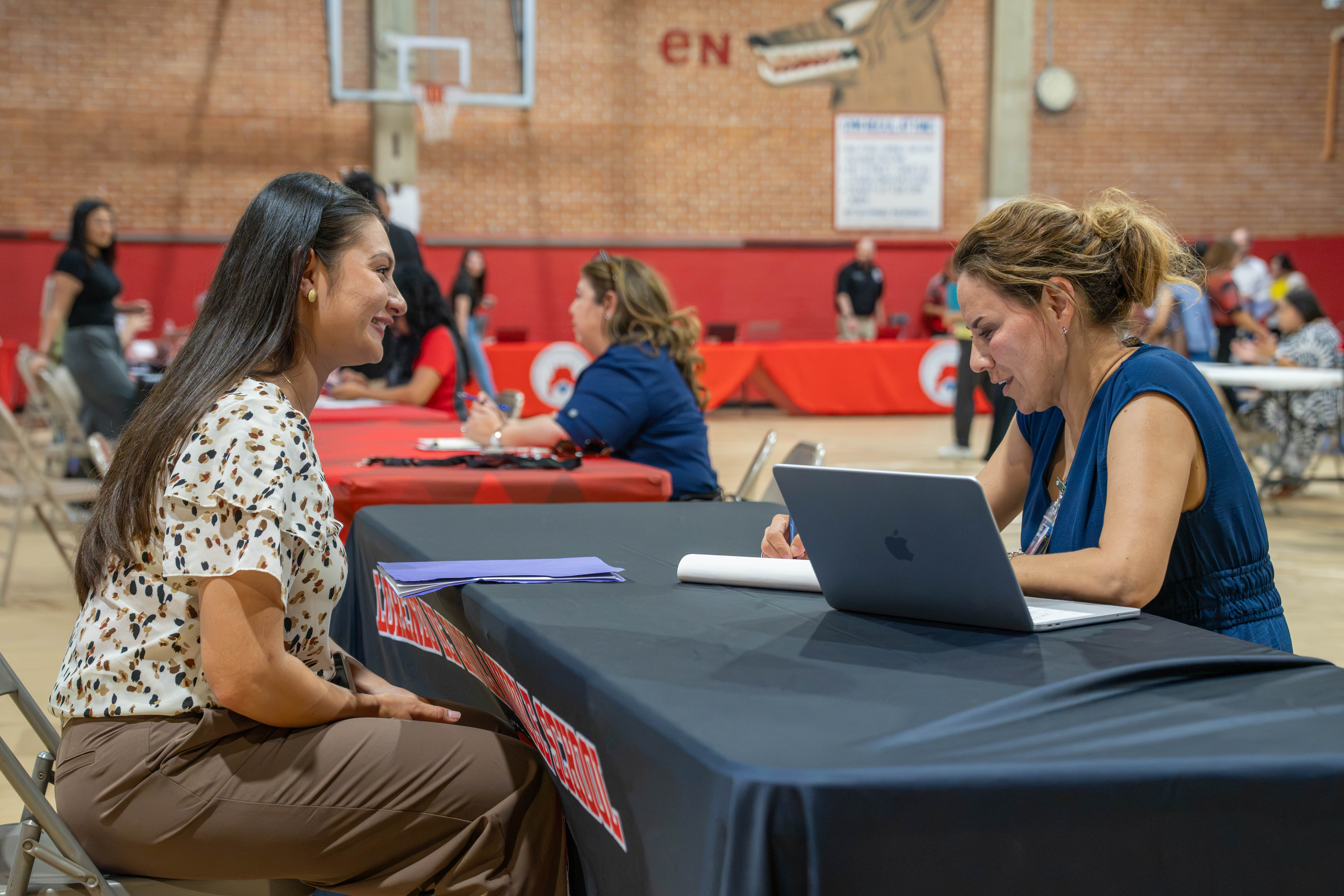 Two women sit at a table covered with a black tablecloth, one using a laptop and the other looking on.