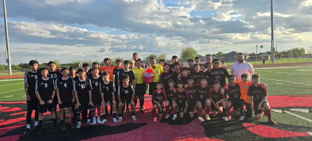 A large soccer team poses for a group photo on a bright green field under a cloudy blue sky.