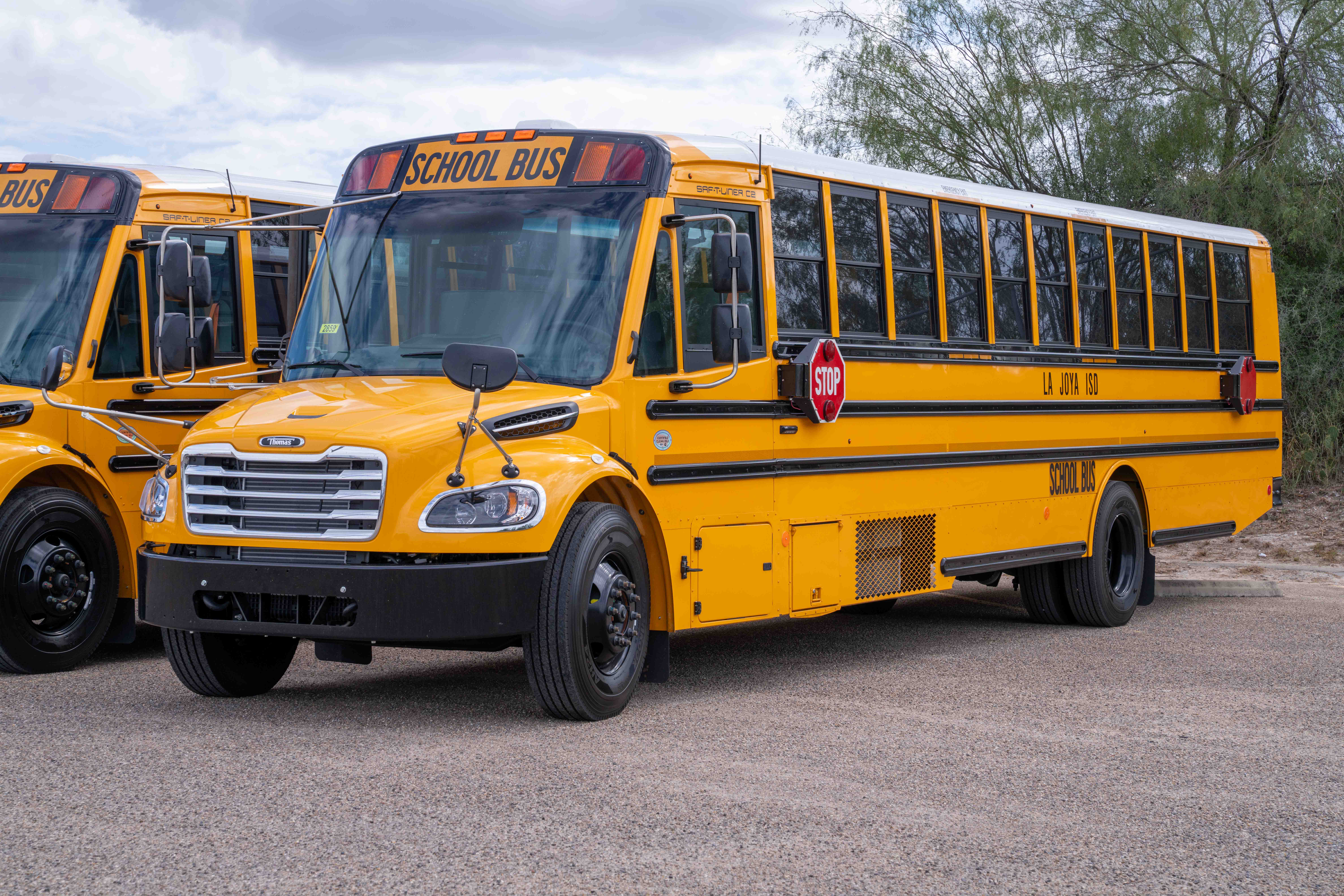 A bright yellow school bus parked on a paved lot under a cloudy sky, with another bus partially visible on the left.