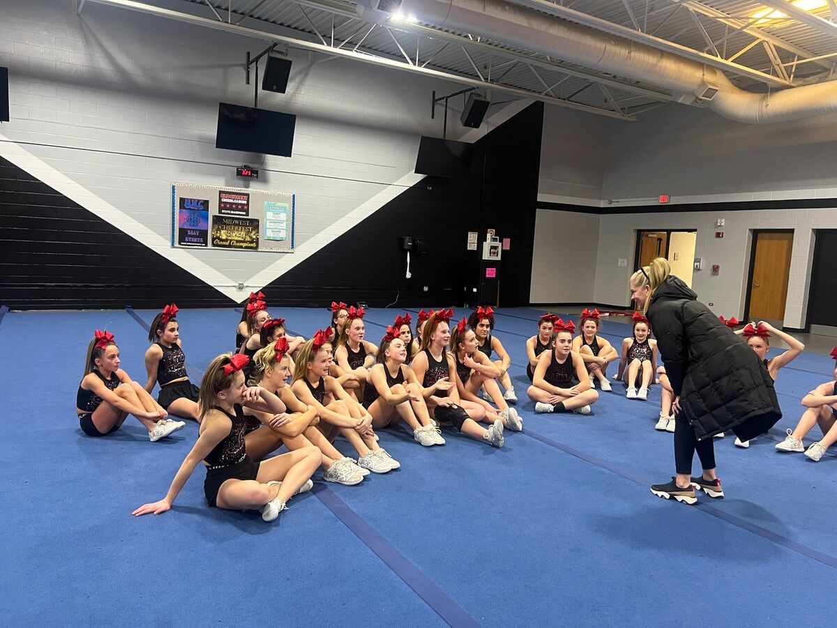 A group of young cheerleaders in black and red uniforms sit on a blue mat, listening to superintendent.