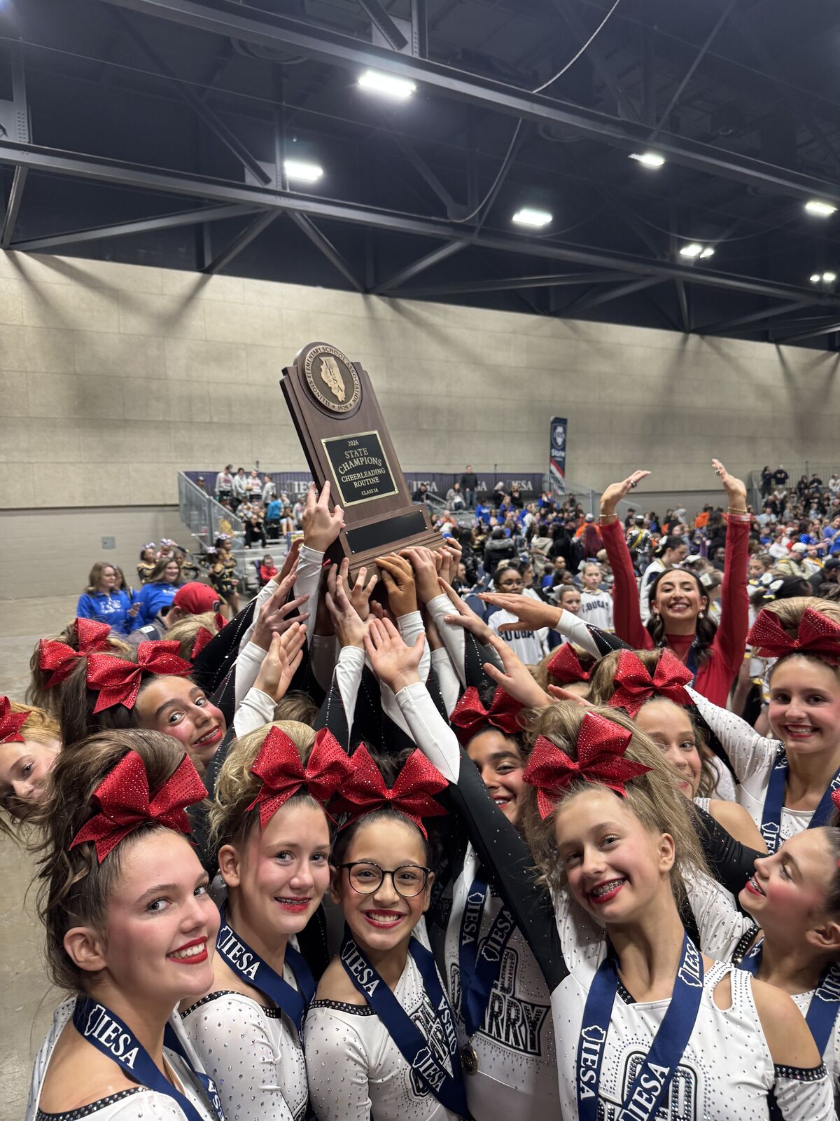 A group of smiling cheerleaders in matching uniforms hold up a large trophy, celebrating their victory.