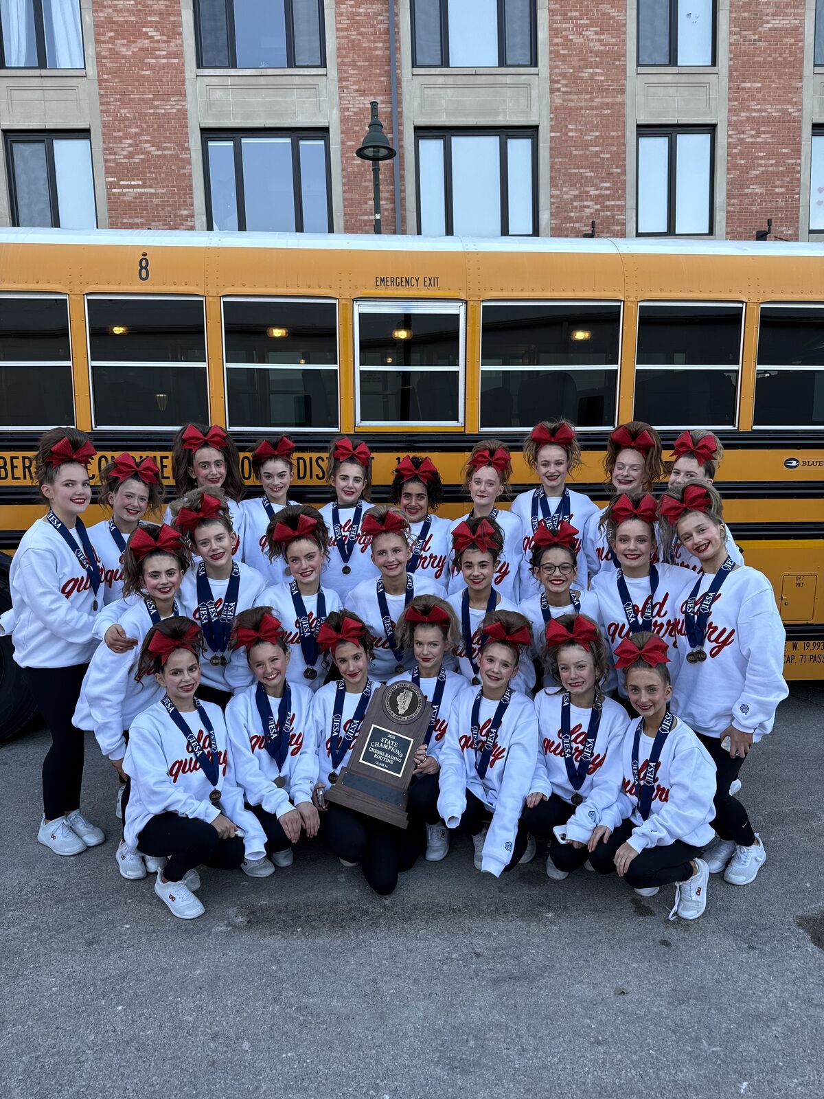 A group of young cheerleaders in white sweatshirts and red bows pose in front of a yellow school bus, holding a trophy.