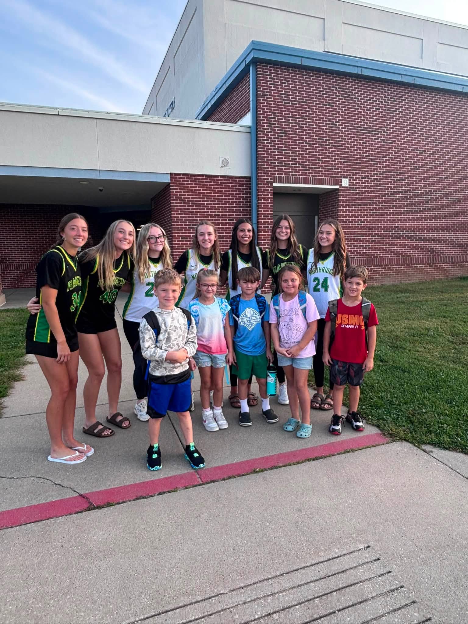 A group of older youths in basketball jerseys stand behind several younger children outside a brick building.