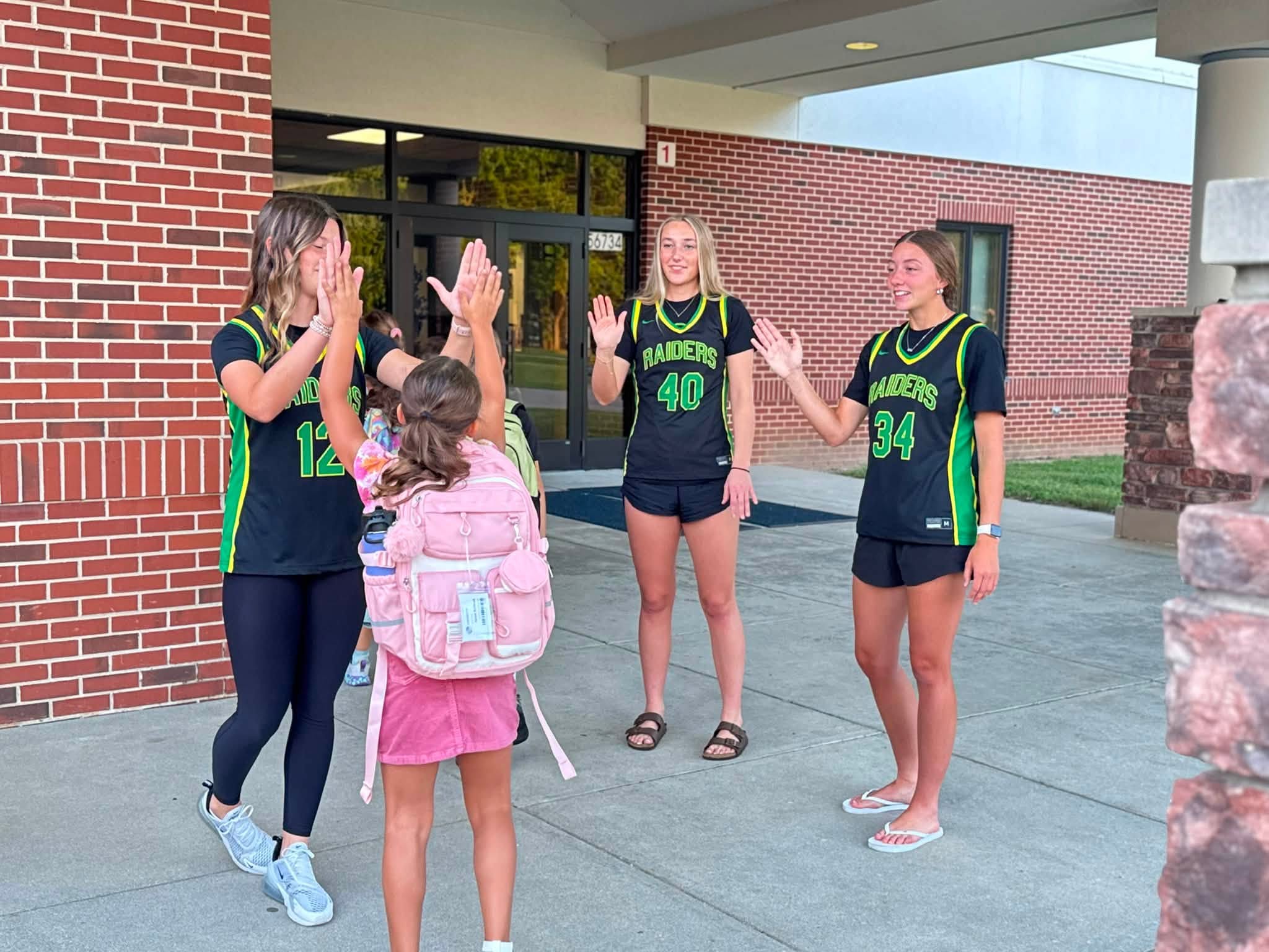 Three high school students in Raiders basketball jerseys high-five a young student wearing a pink backpack outside a school entrance.