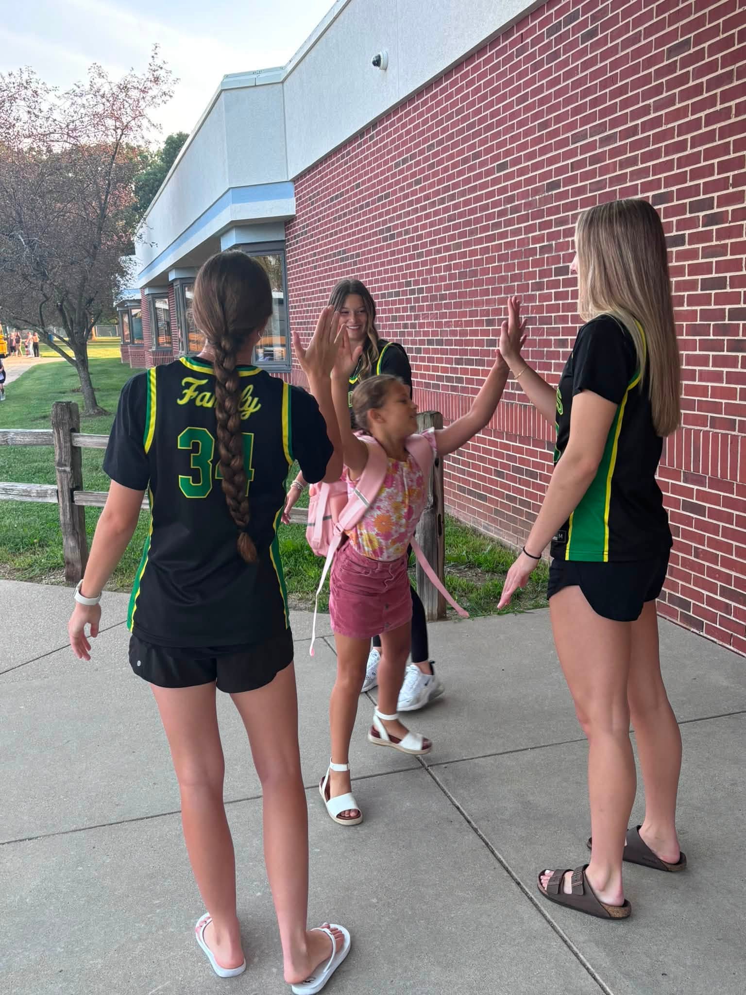 A young girl in a pink backpack high-fives two female athletes wearing black and green jerseys outside a brick building.