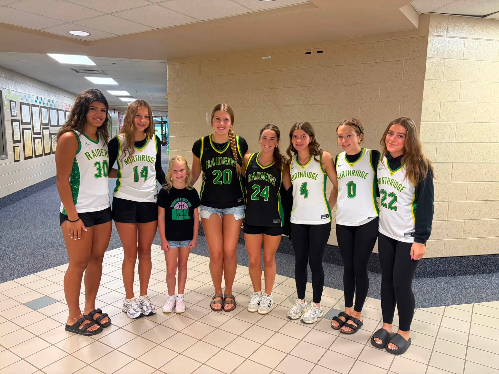 Seven female basketball players and a young child stand together in a school hallway, wearing green and white jerseys that read 'NORTHRIDGE' and 'RAIDERS'.