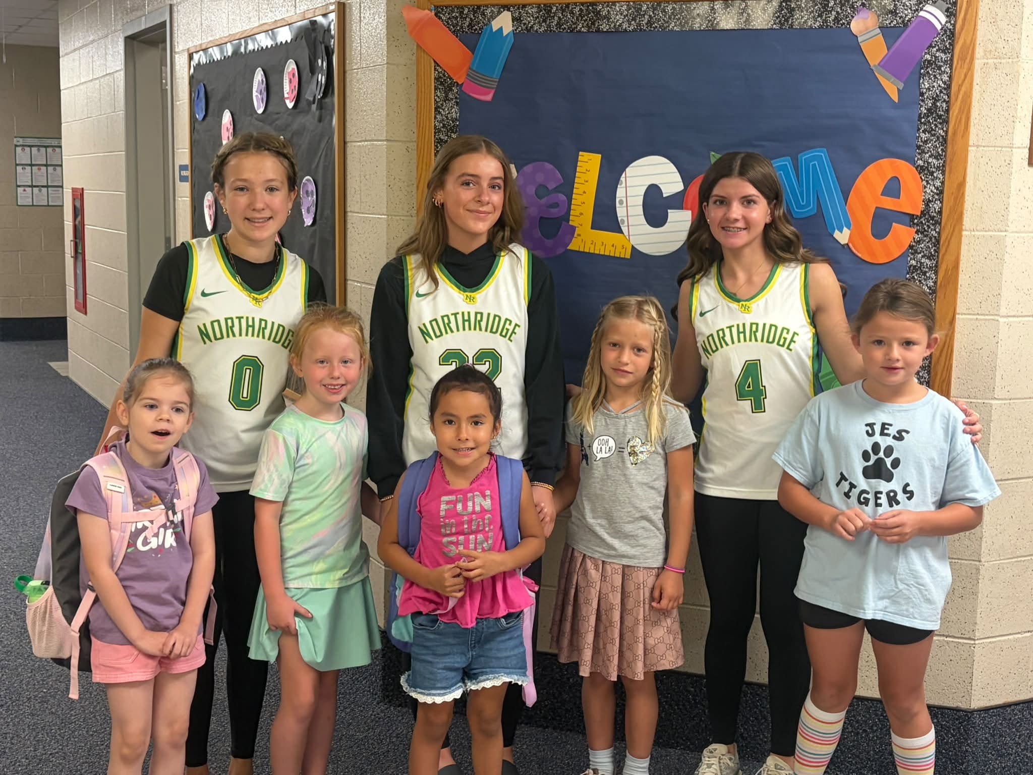 Three older students in Northridge basketball jerseys pose with six younger children in a school hallway.