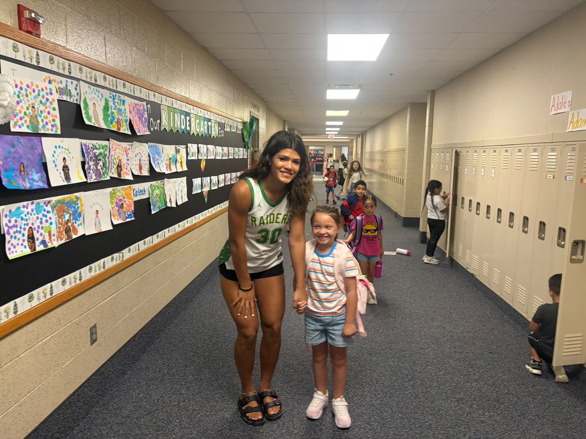 A tall athlete wearing a green and white Raiders jersey poses with a smiling young student in a school hallway.
