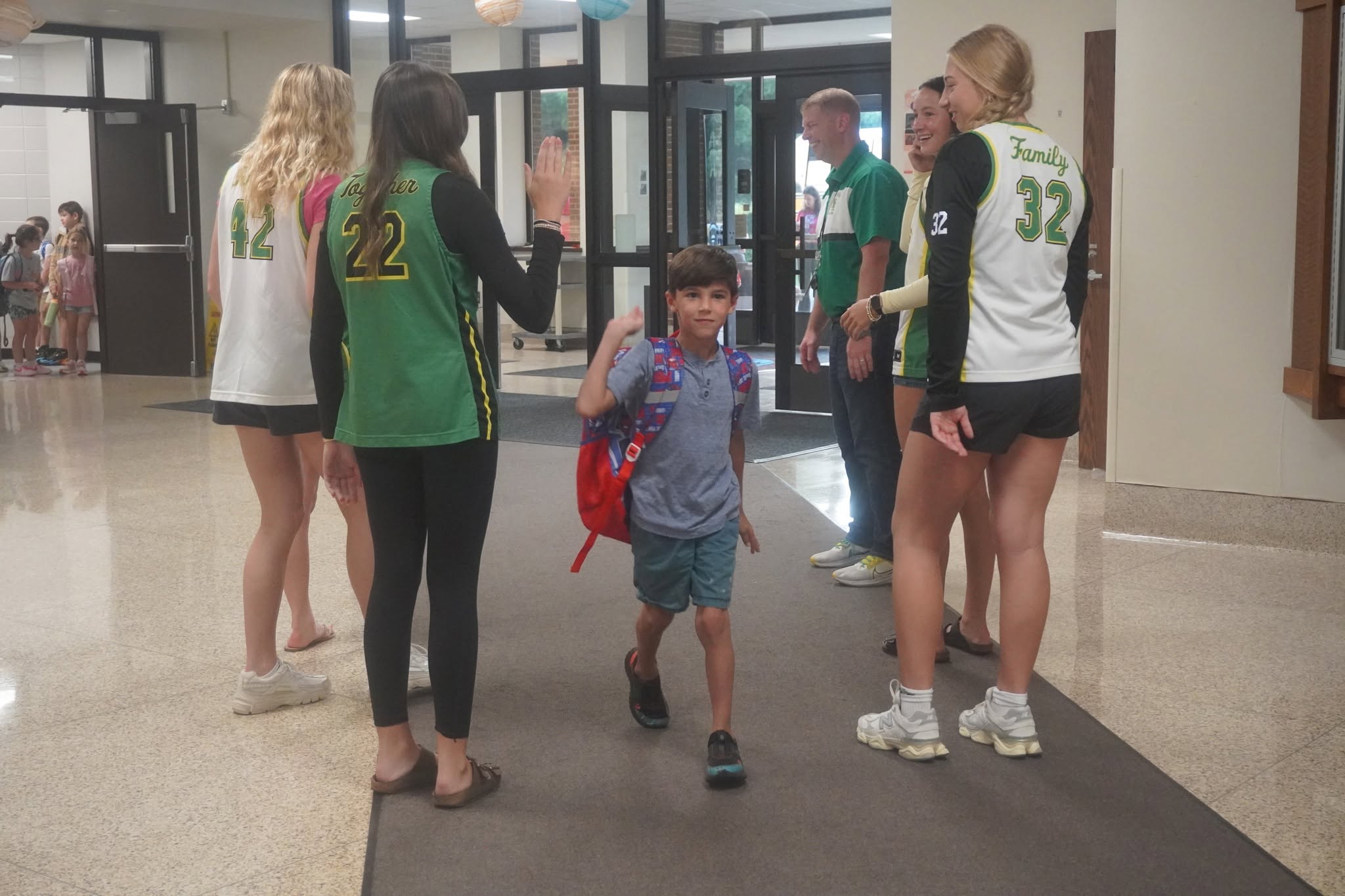 A young boy with a red backpack walks down a school hallway, high-fiving older students wearing matching sports jerseys.