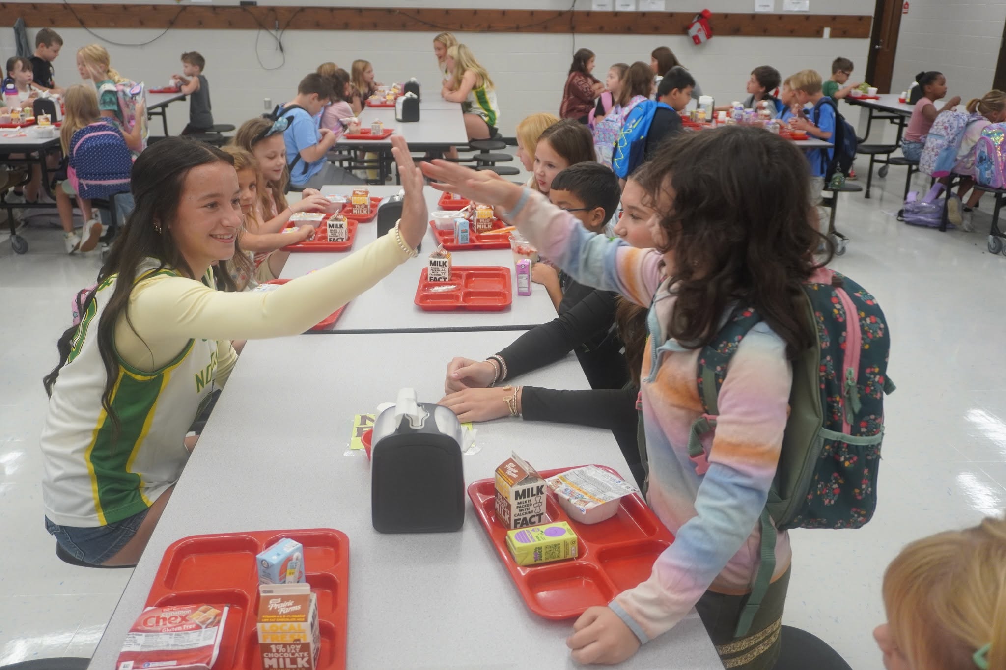 Two girls share a cheerful high-five across a lunch table in a busy school cafeteria.