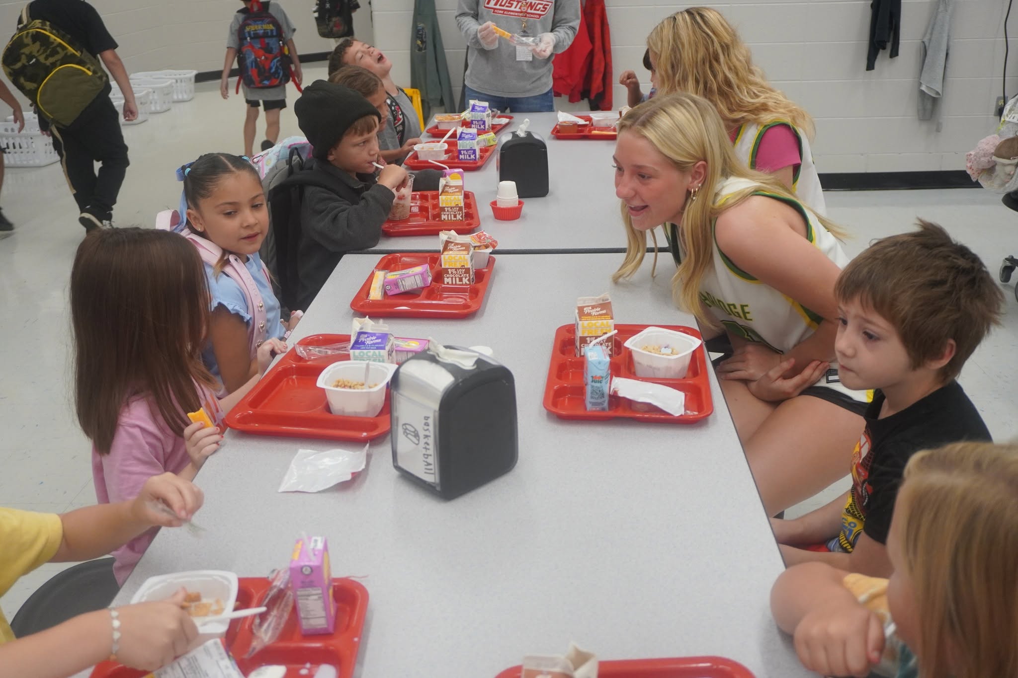 Students sit at long cafeteria tables during lunch, with red trays holding cartons of milk and food.