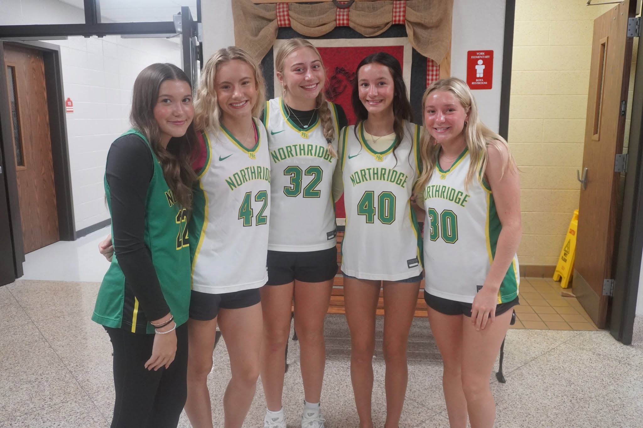 Five young women wearing Northridge basketball jerseys pose together indoors, smiling at the camera.