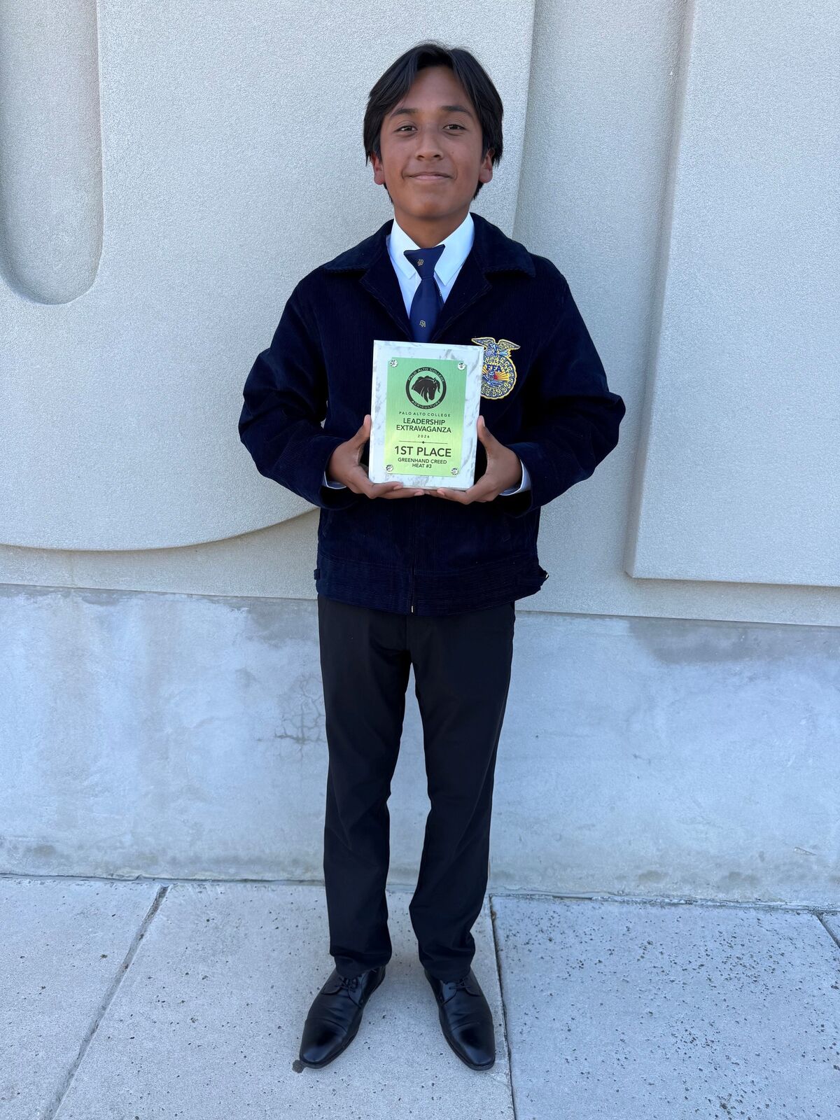 A young man in a dark blue jacket and tie proudly holds a first-place award plaque outdoors.