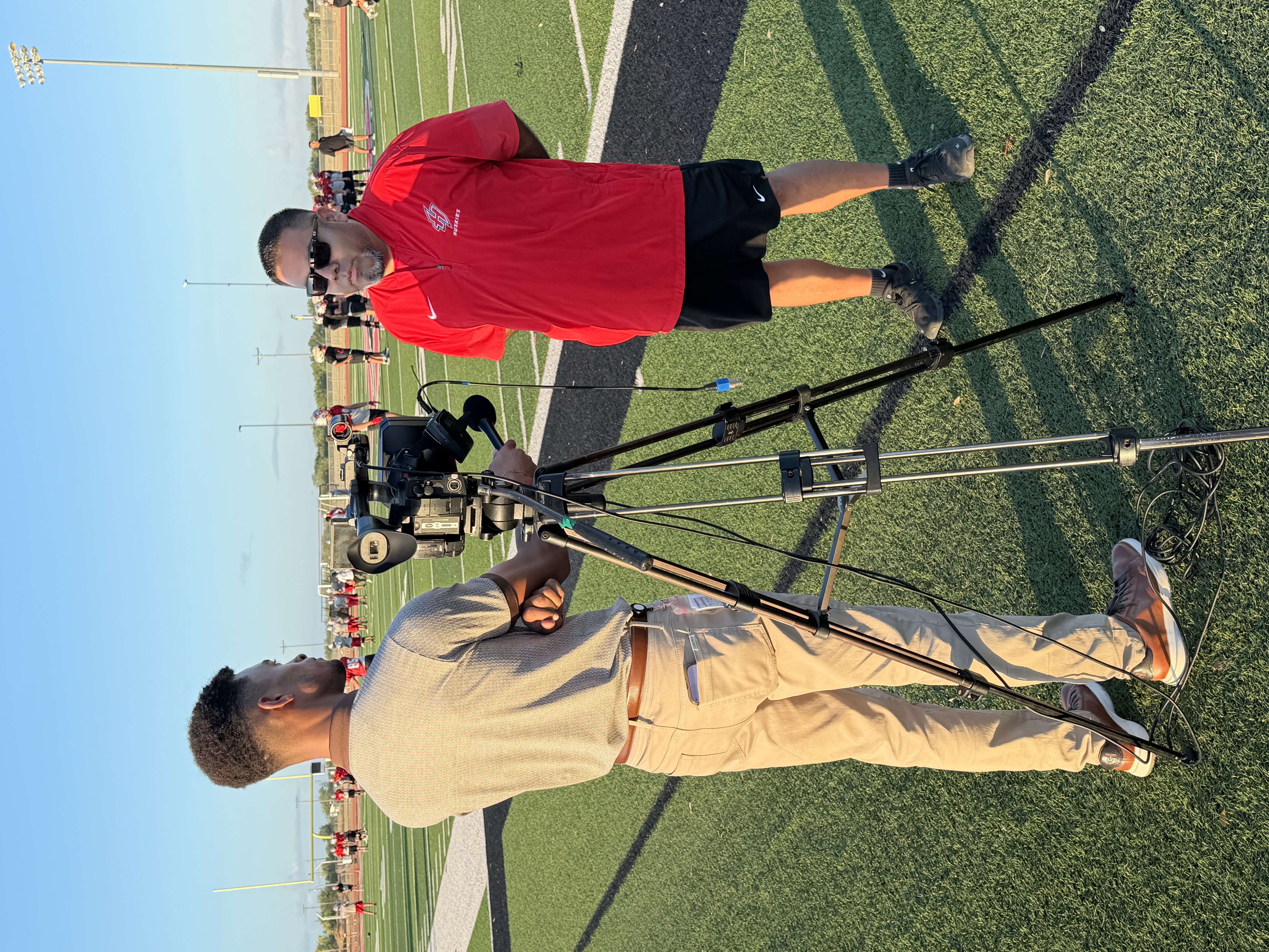 A videographer operates a camera on a tripod at an outdoor sports field, interviewing a coach in a red shirt.