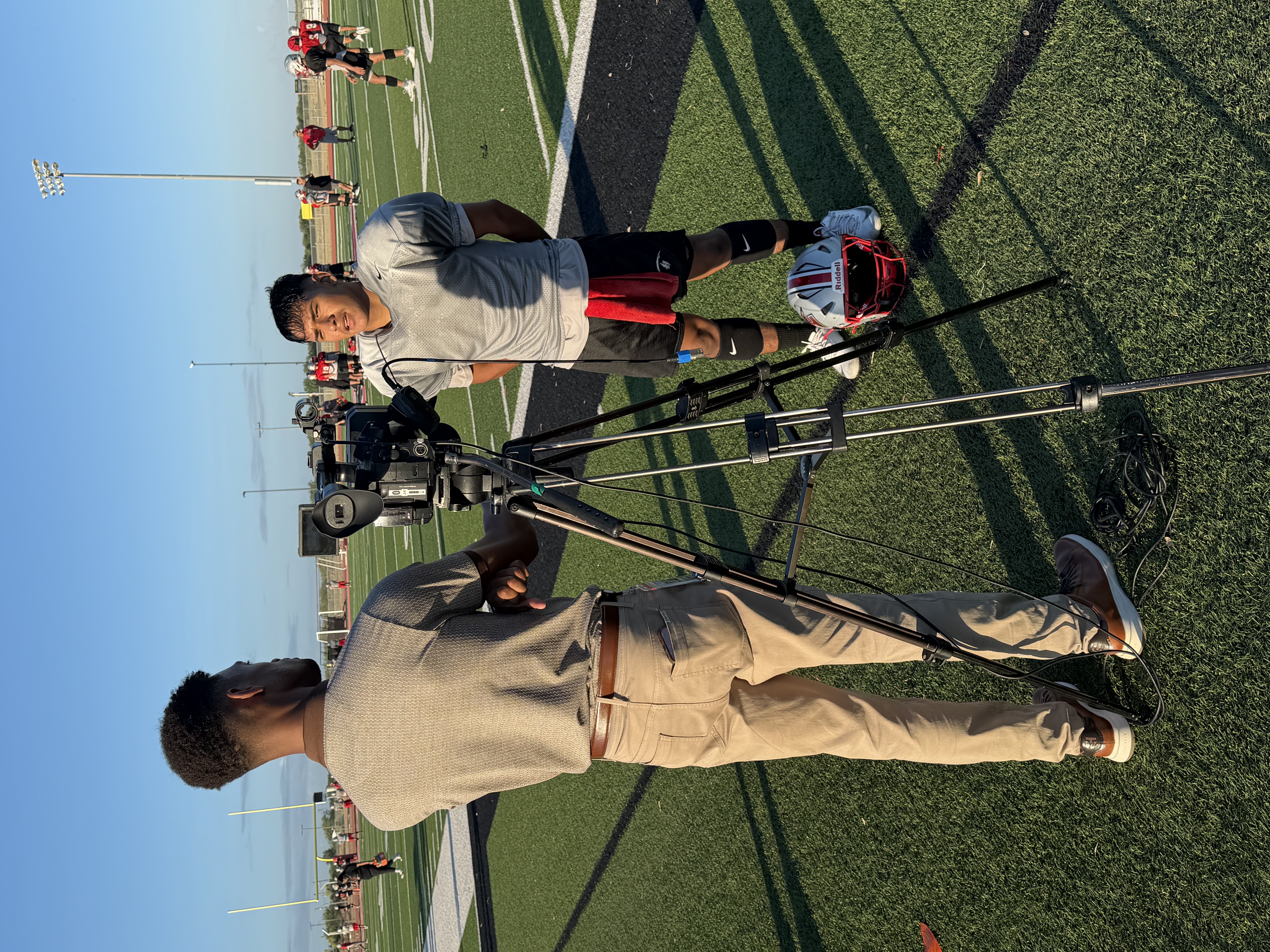 A cameraperson stands behind a tripod-mounted camera, filming an athlete on a sunny football field.