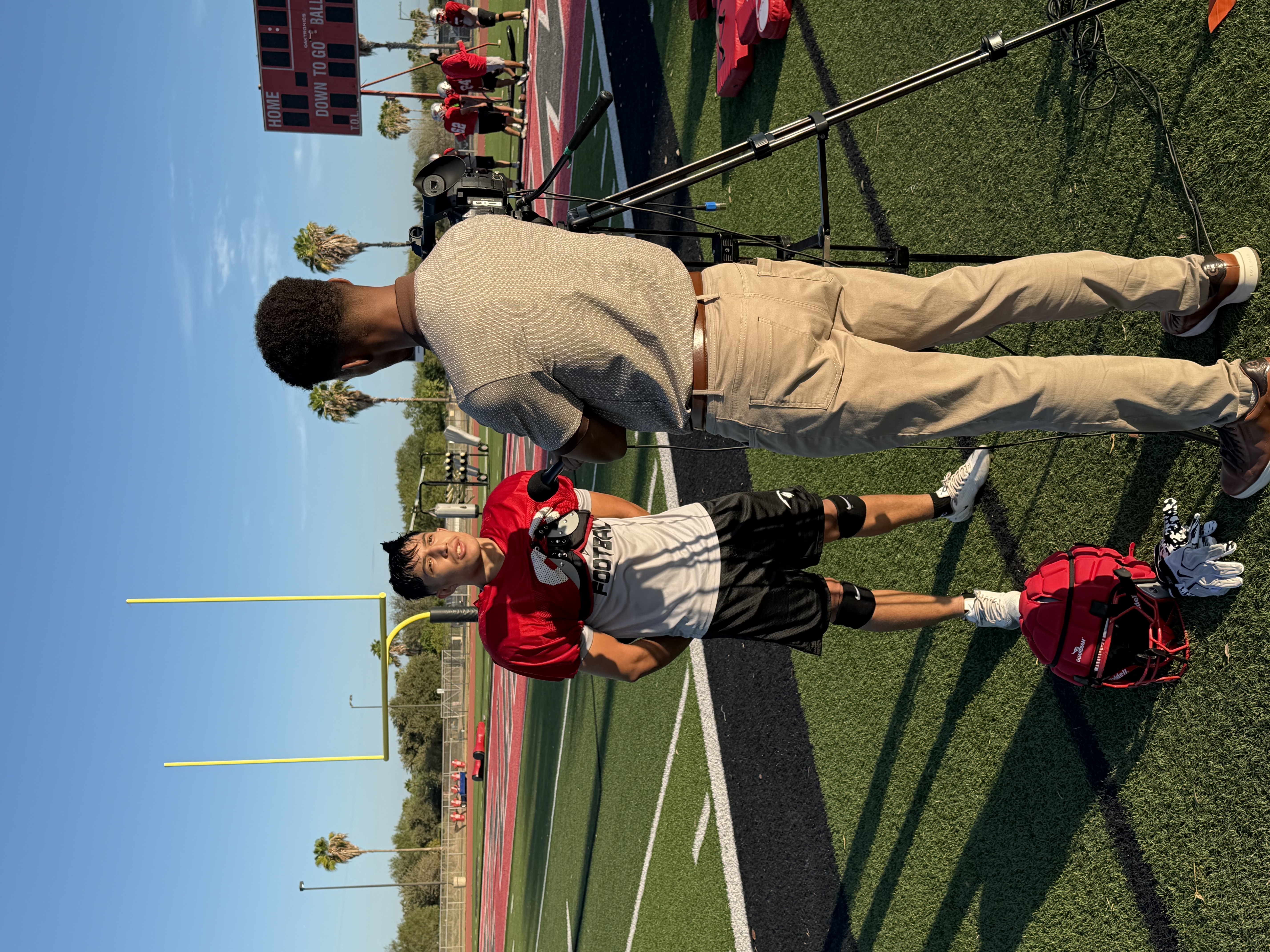 A camera operator films a football player on a sunny outdoor field during practice.