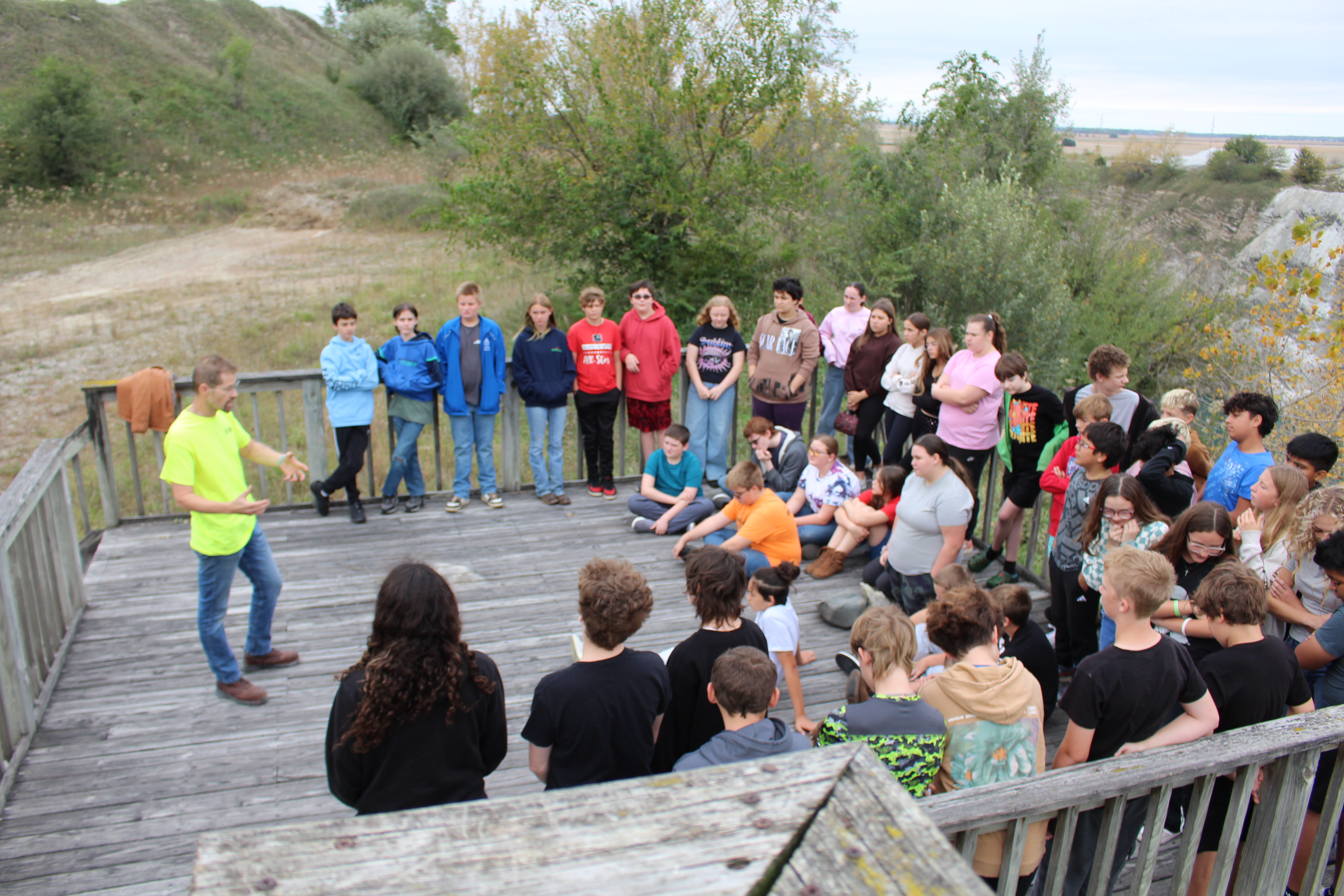 A group of students stands and sits on a wooden observation deck listening to an instructor outdoors.