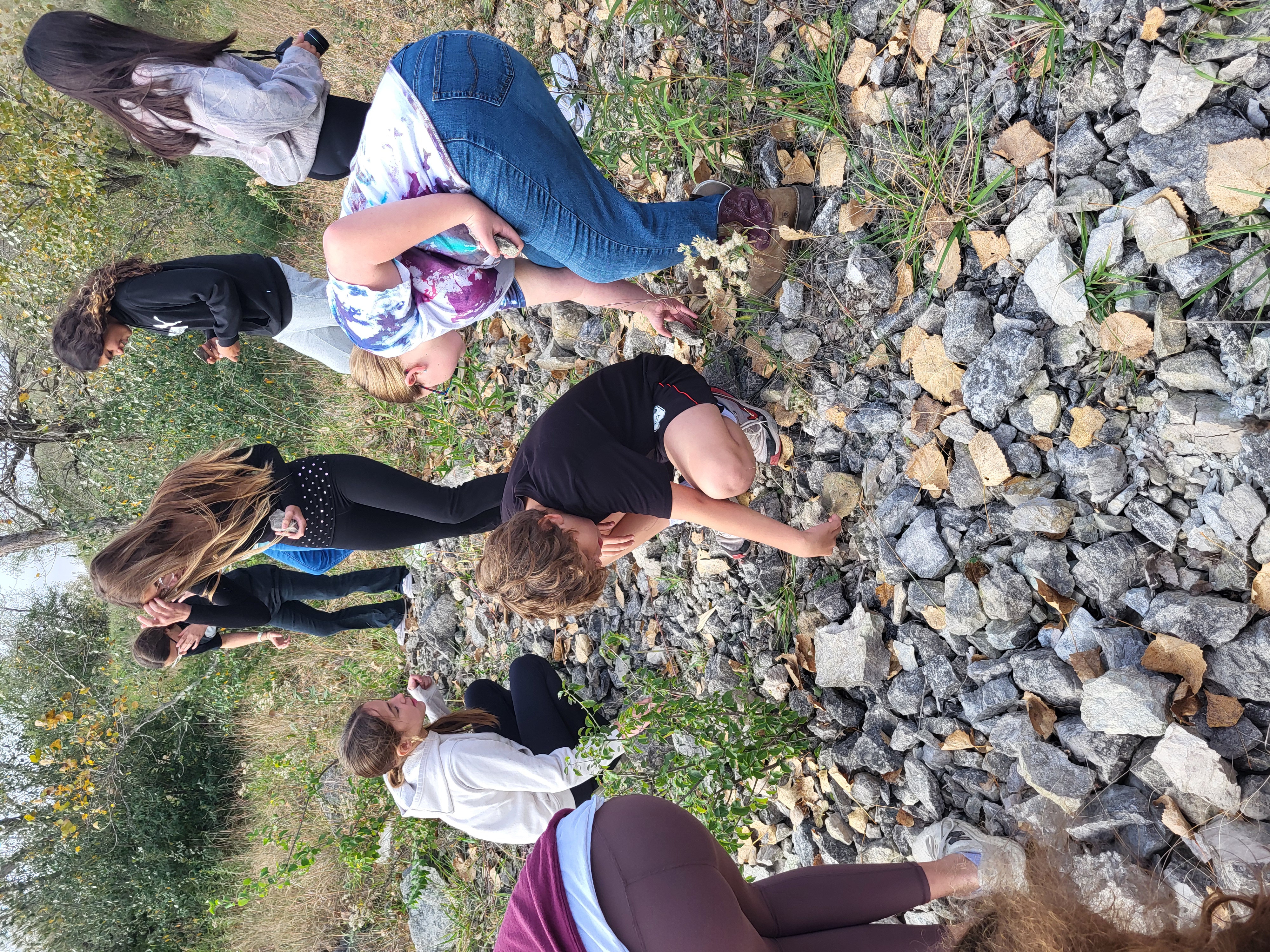A group of people explore an outdoor rocky area, bending down to examine plants and rocks among fallen leaves.