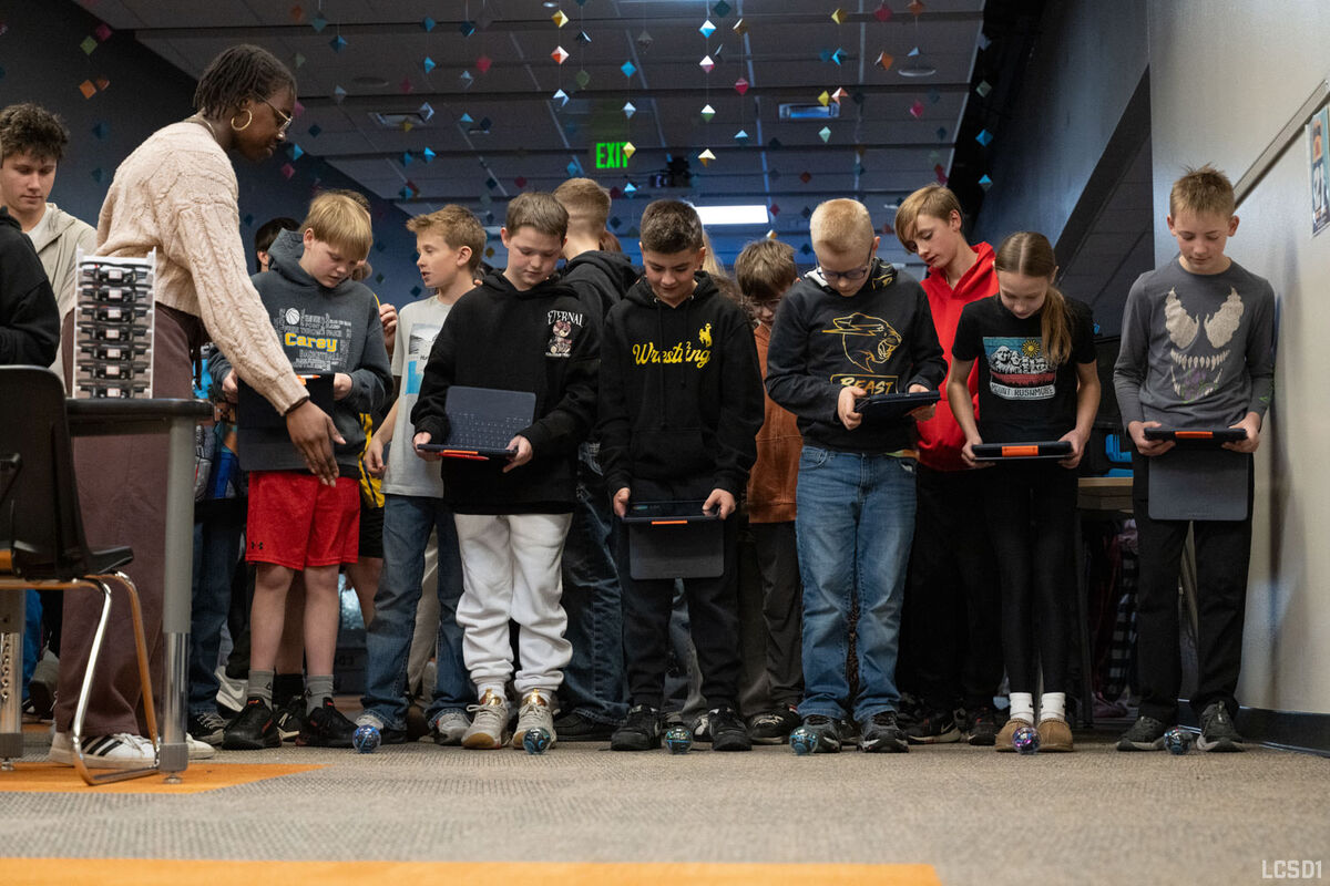 A teacher guides students as they interact with small robots and tablets in a classroom.