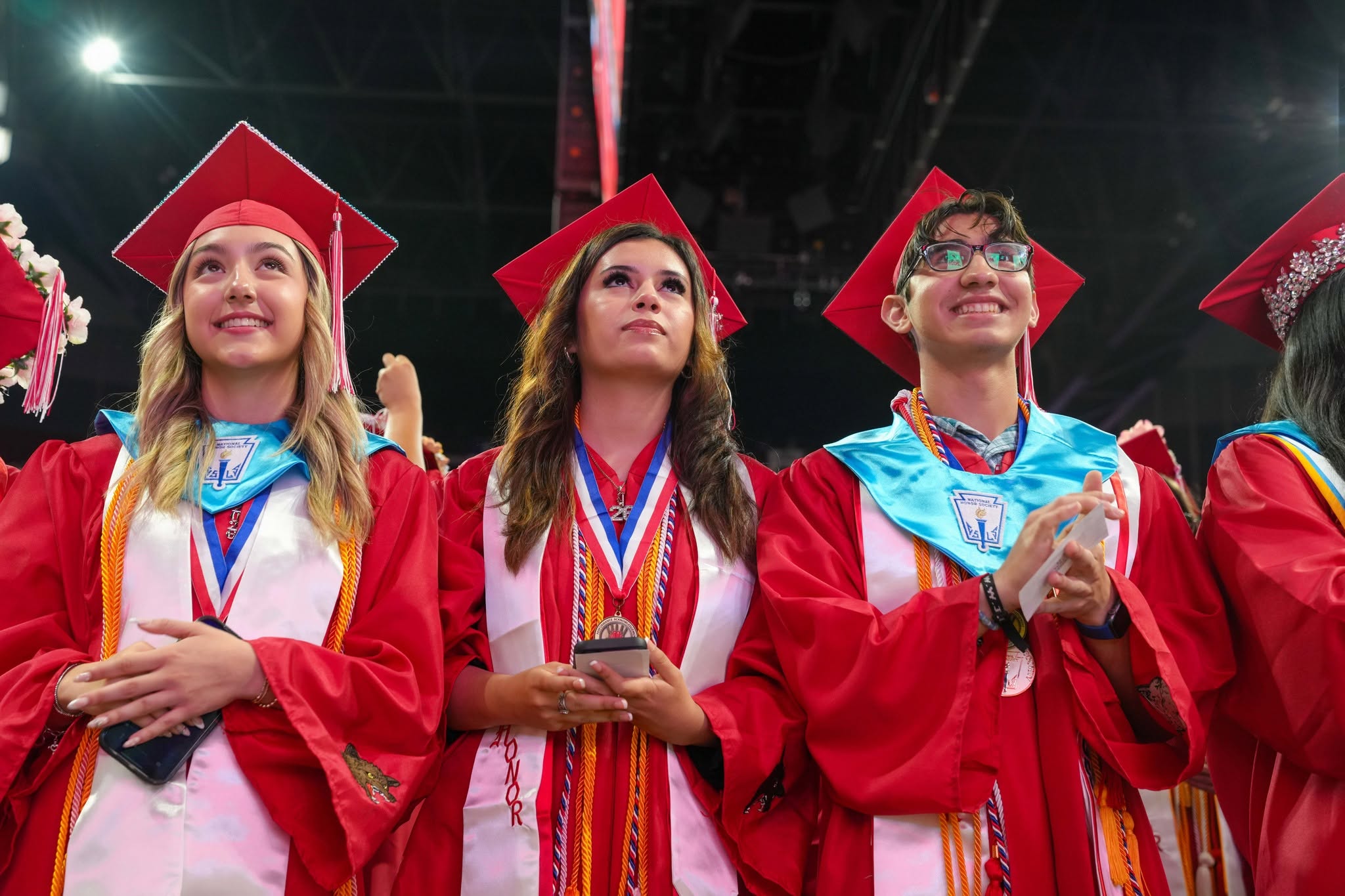 Three young graduates in red caps and gowns look forward, smiling and holding phones during a ceremony.