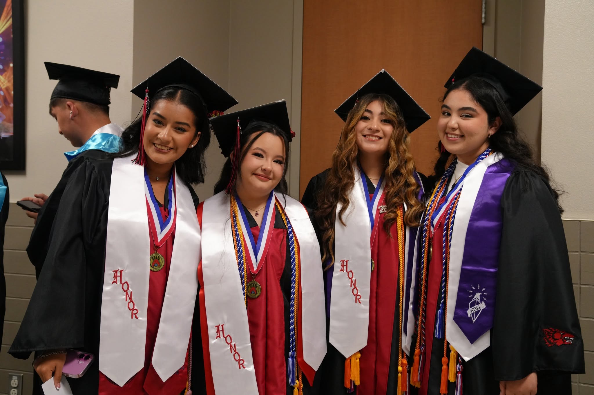 Four smiling graduates wearing caps, gowns, and honor stoles pose together indoors.