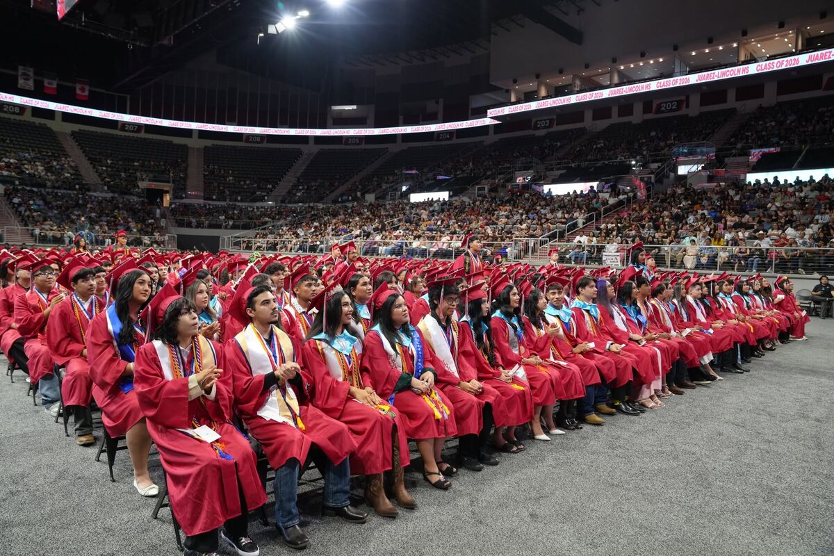 A large group of high school graduates in red caps and gowns sit seated on chairs inside a packed arena.