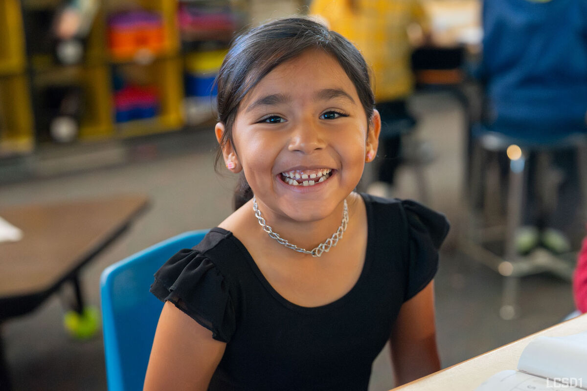 A young girl with dark hair smiles broadly, wearing a black shirt and a silver necklace.