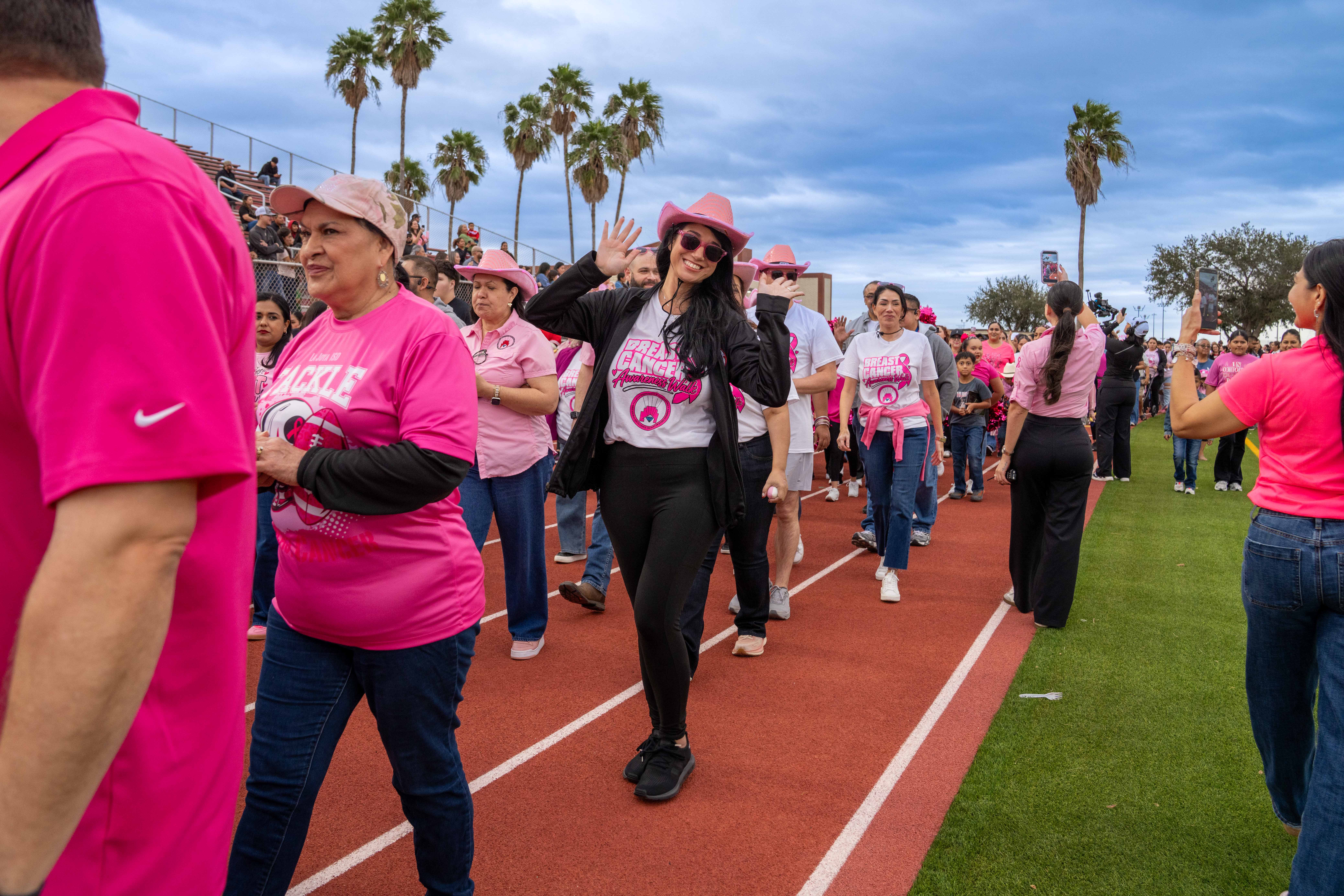 A group of people wearing pink shirts and hats walk on a red running track during a breast cancer awareness event.