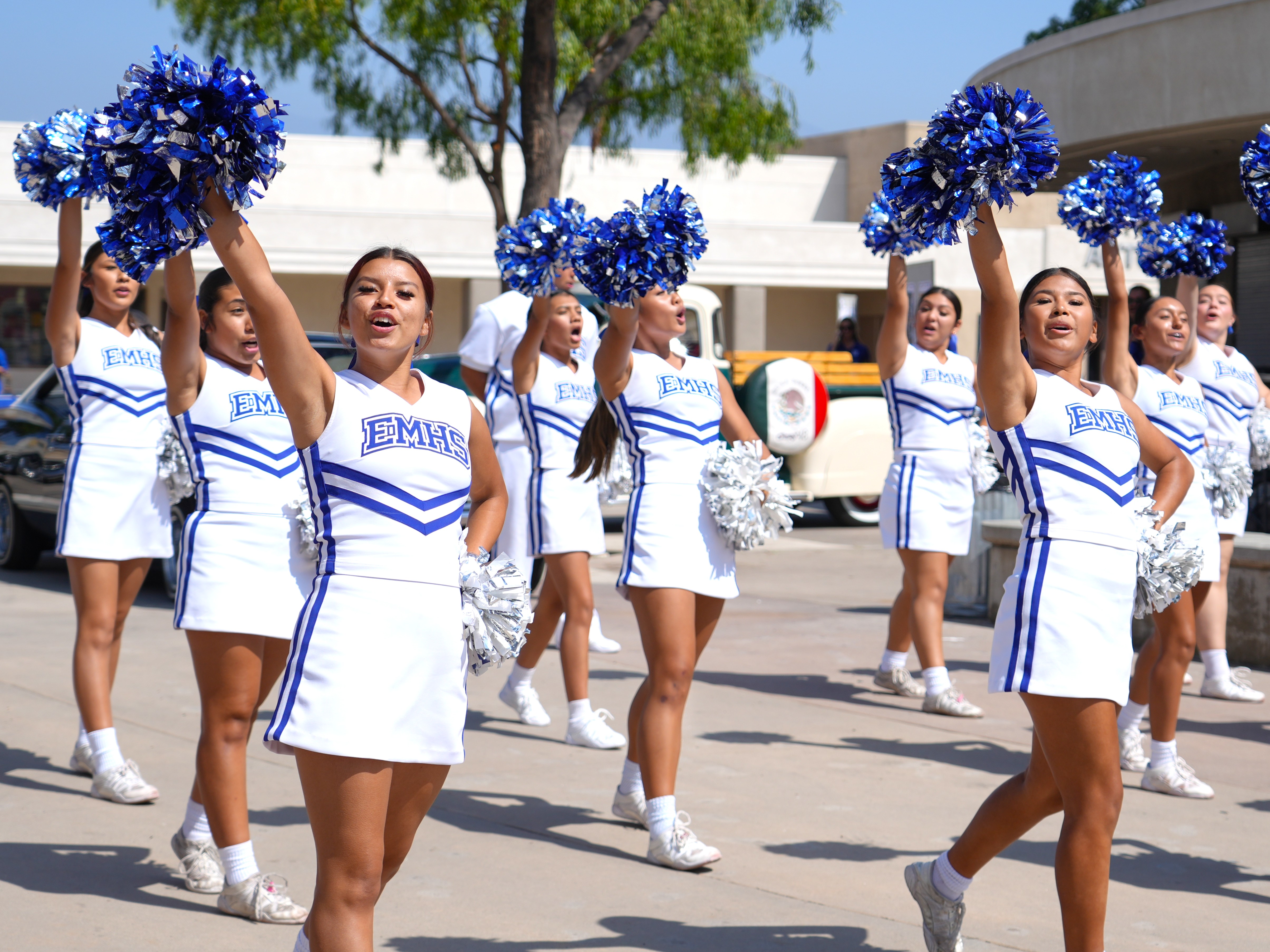 A line of high school cheerleaders in white and blue uniforms with the text EMHS holds up blue pom-poms while performing outside.