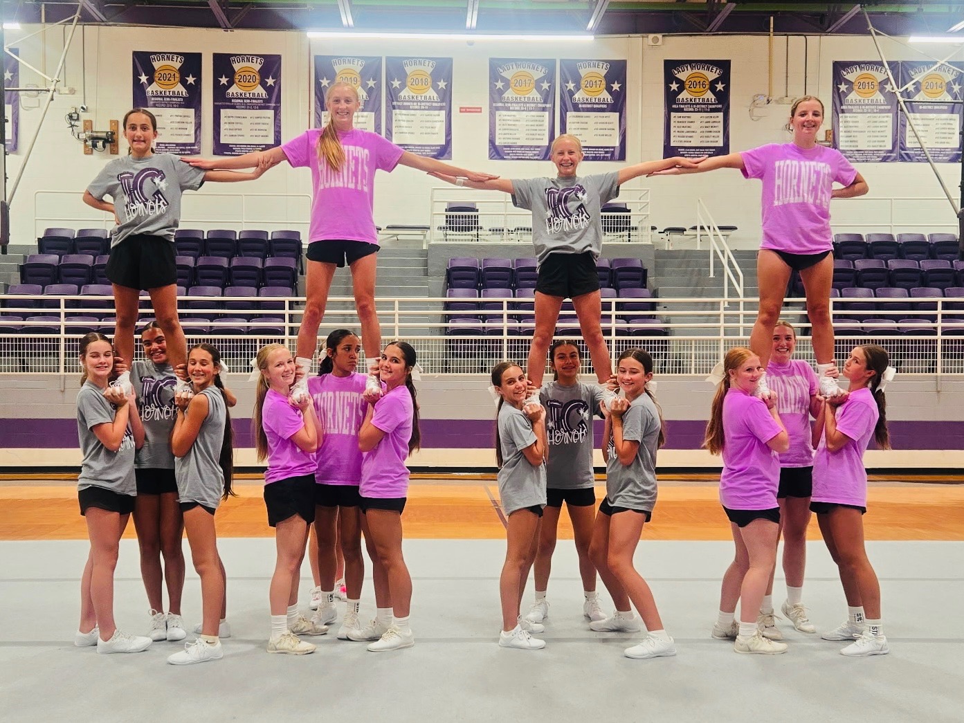 A cheerleading squad performs a pyramid formation in a gymnasium, with some members standing on others' shoulders.