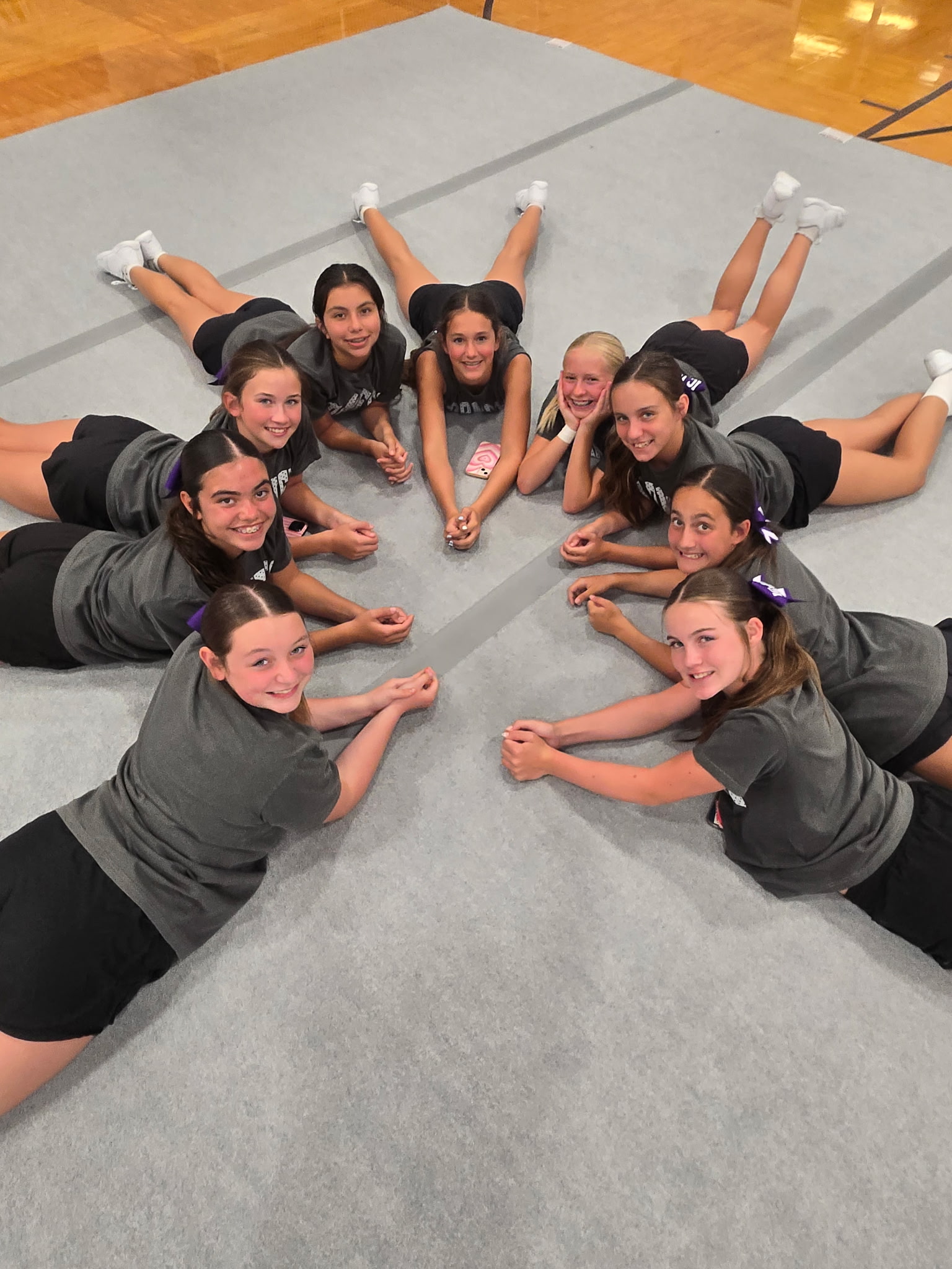 A group of young cheerleaders lie on a mat in a circle, smiling at the camera.