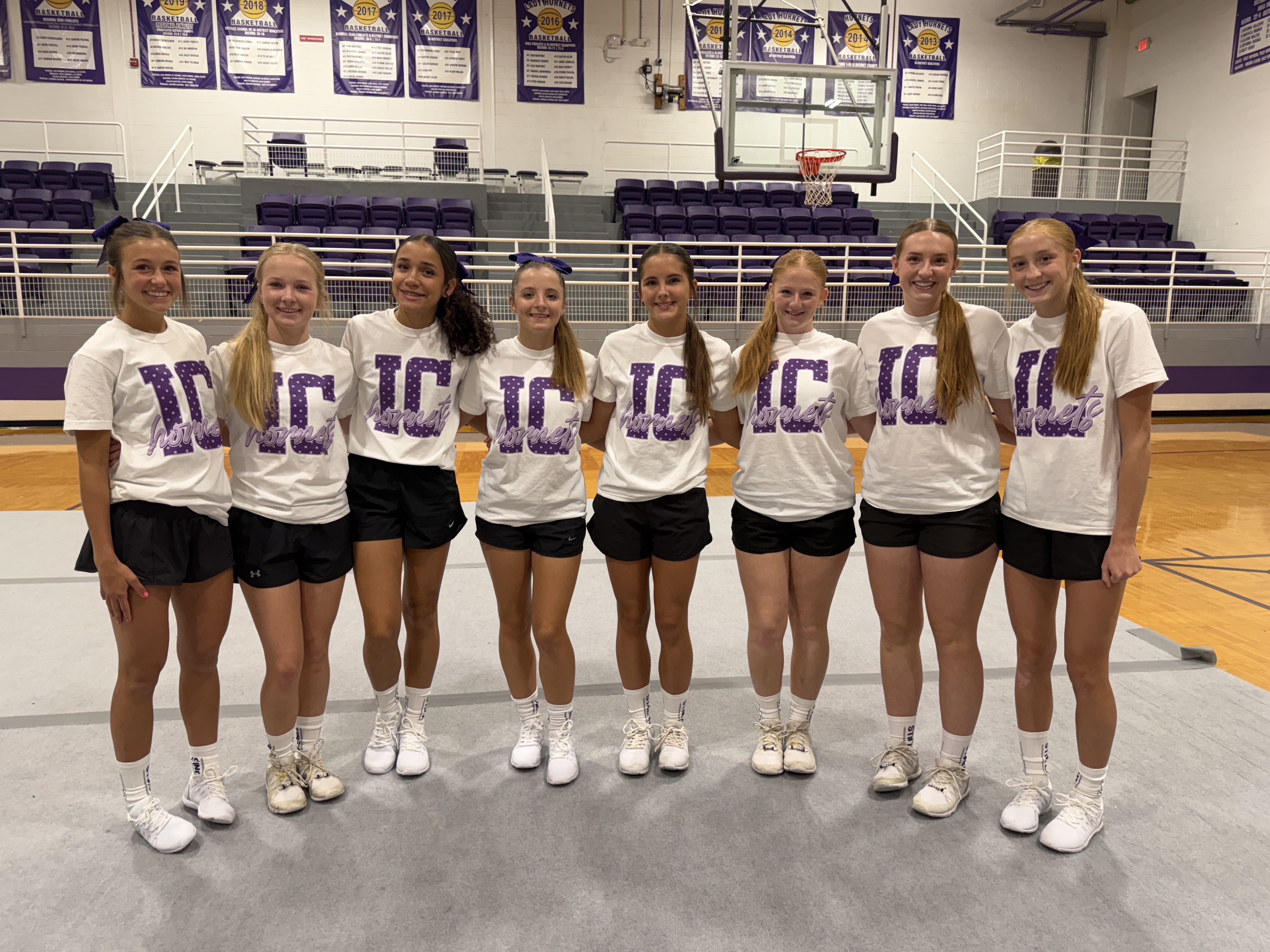 Eight young women in matching white t-shirts with purple lettering stand together in a gymnasium.