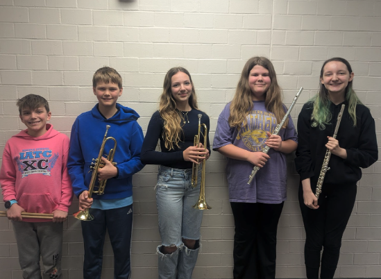 Five young musicians stand in a line, holding drum sticks, brass instruments, and woodwind instruments.