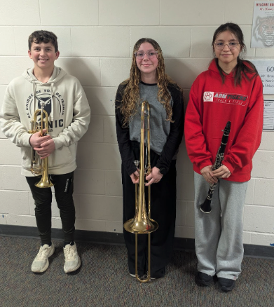 Three young musicians stand together, holding a trumpet, a trombone, and a clarinet.