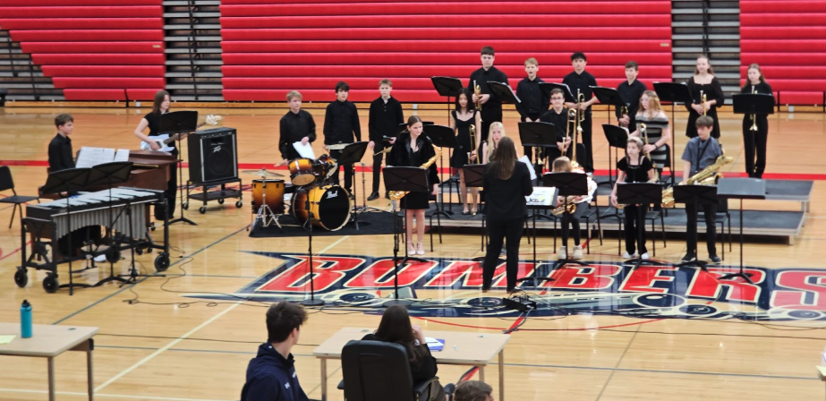 A school band performs on a gymnasium floor, with students playing various instruments like saxophones, trumpets, and drums.