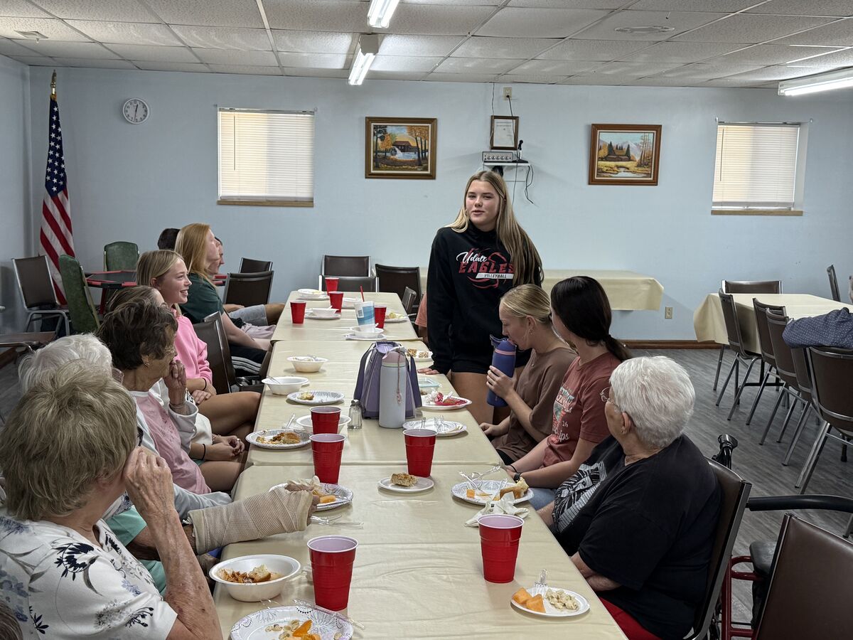 A group of people, including seniors and young women, are gathered around a long table set for a meal.