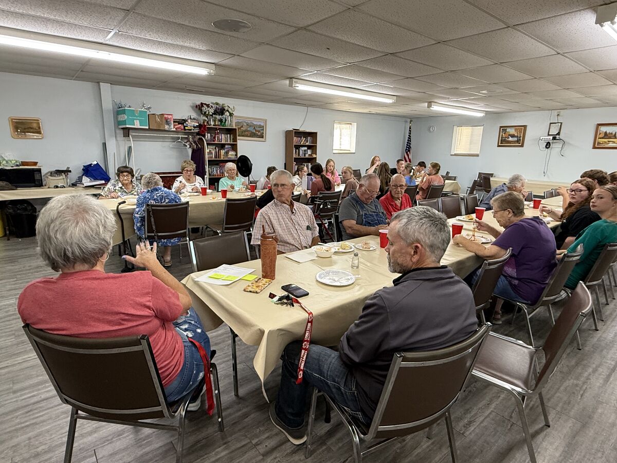 A diverse group of people of various ages are seated at tables, enjoying a meal together indoors.