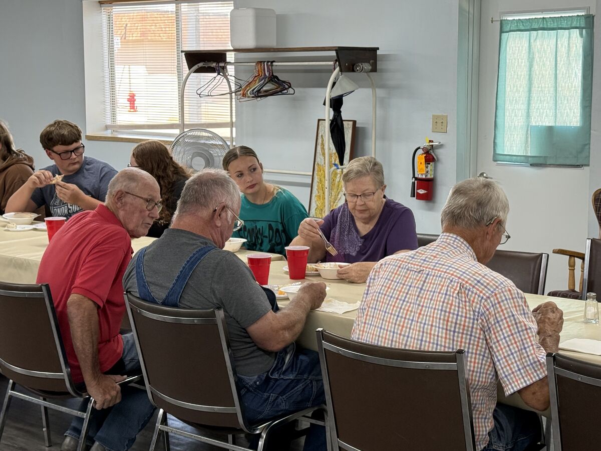 A group of people of various ages are seated around a long table, eating a meal together.