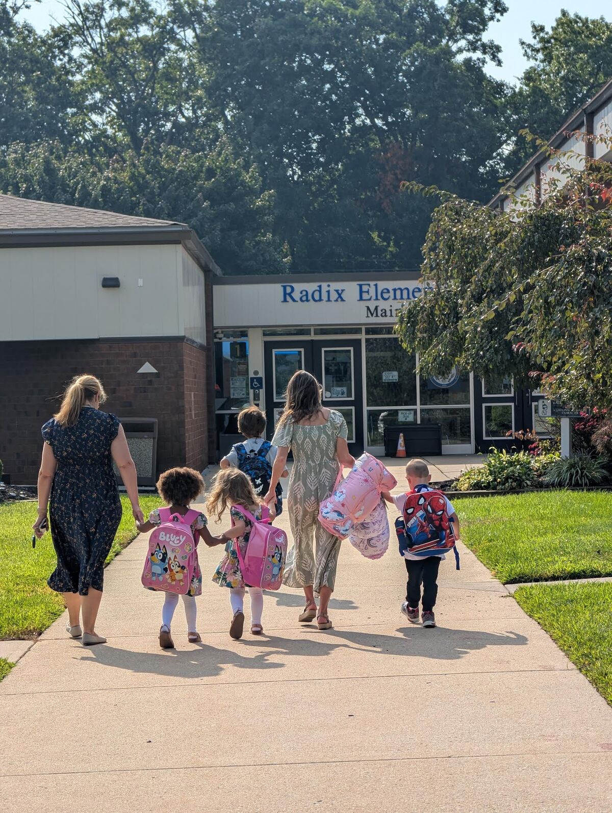 Adults and children walk hand-in-hand along a sidewalk toward the entrance of Radix Elementary School.