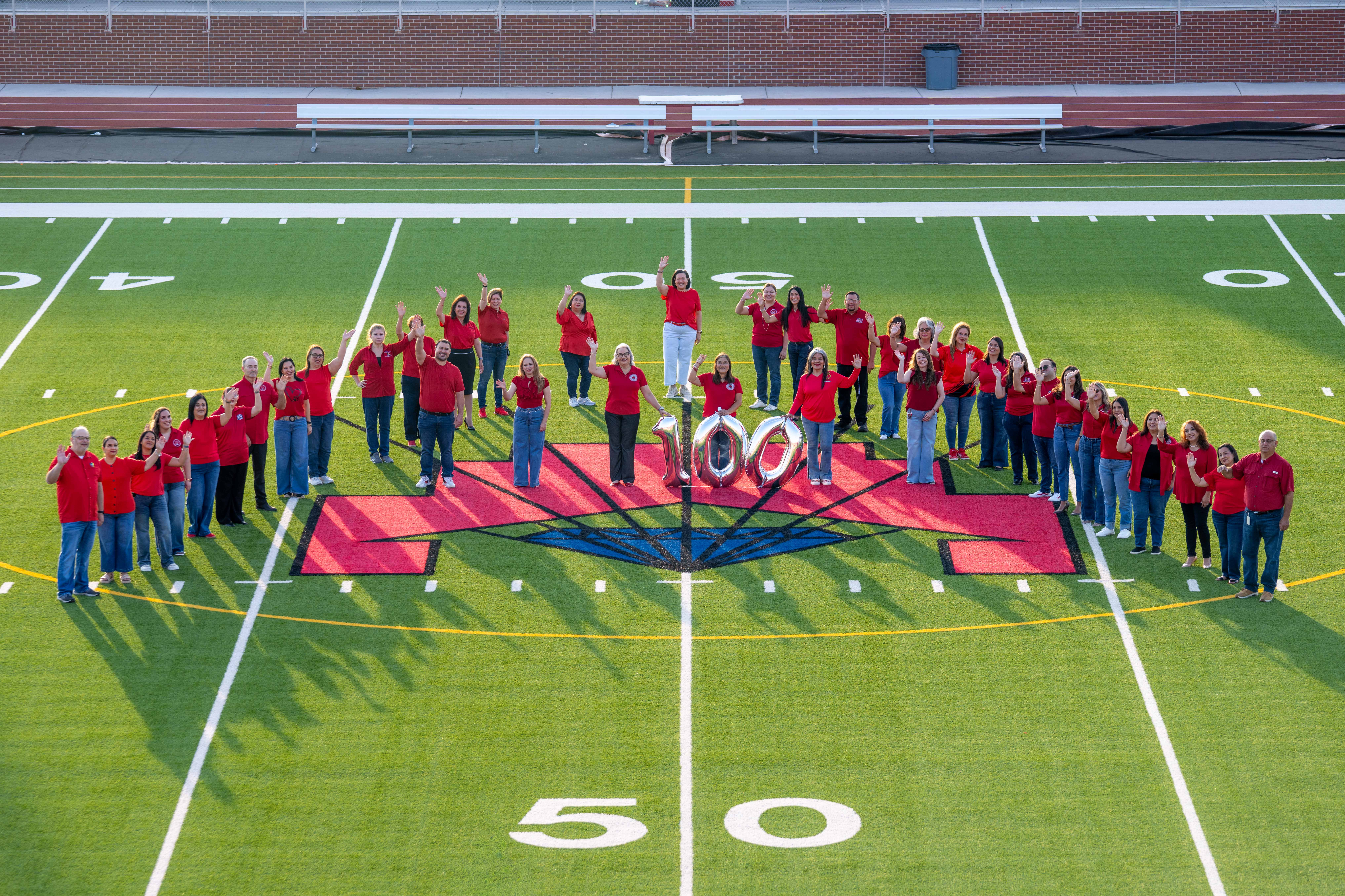 A large group of people dressed in red stand on a football field, posing around giant silver balloons shaped like the number 100.