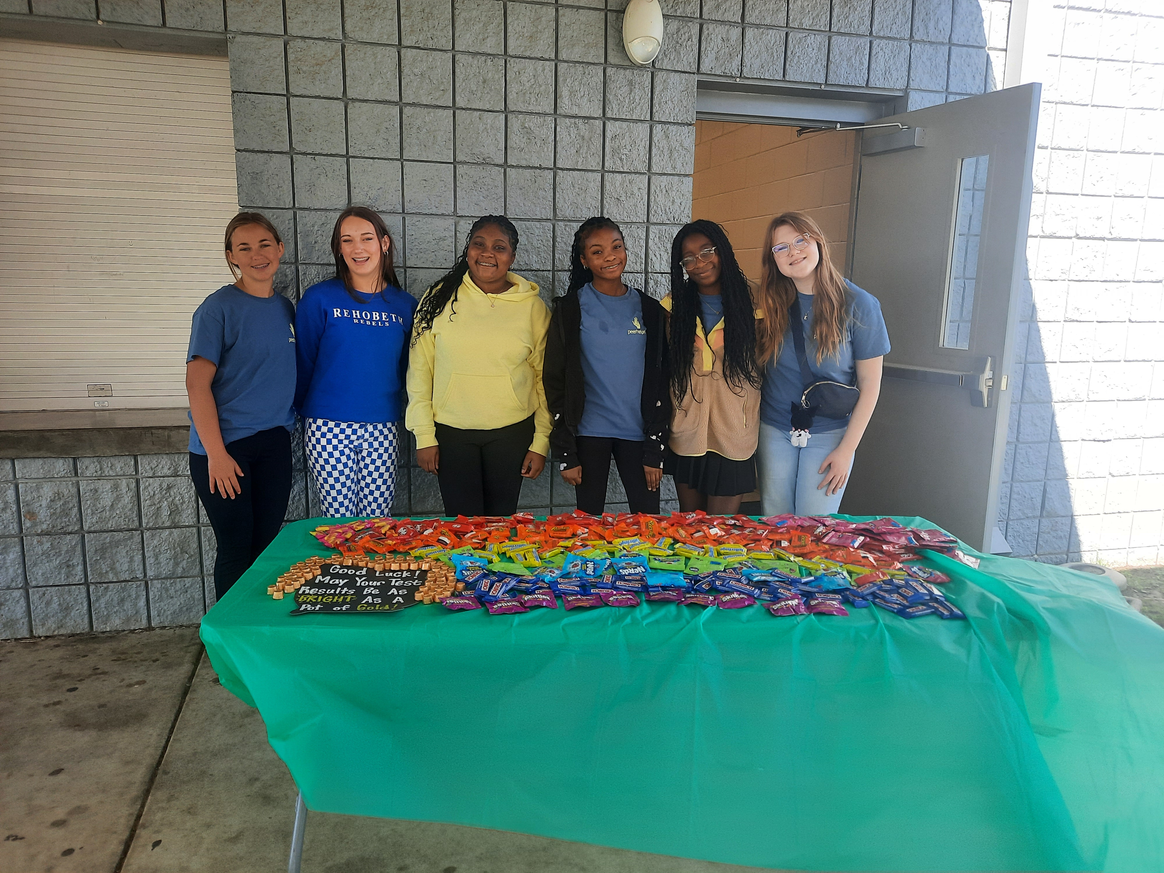 Six smiling students stand behind a table covered in a green cloth, displaying a large assortment of candy.